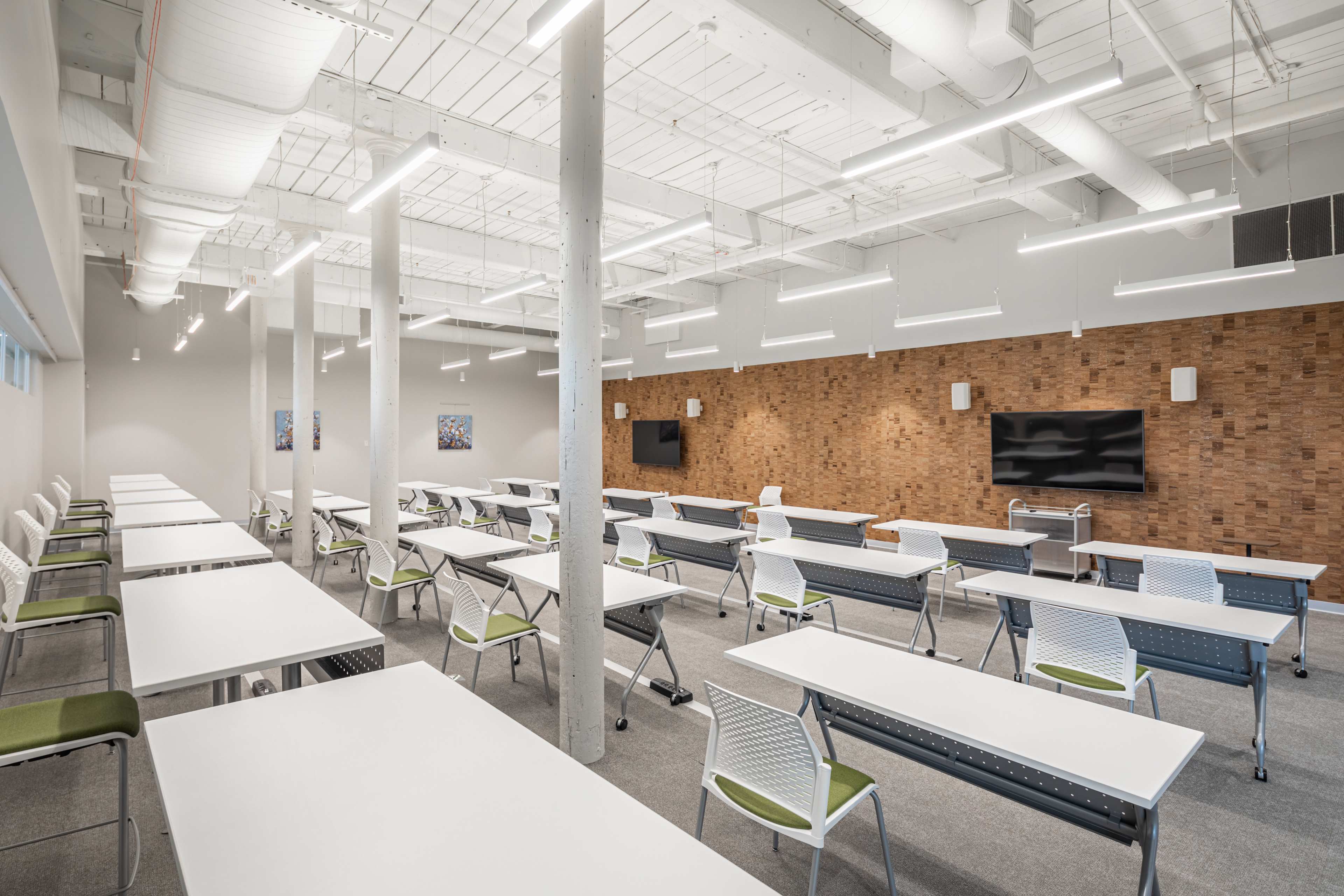 A brightly lit classroom features several rows of folding tables and chairs, with a large television displayed on a wooden accent wall.
