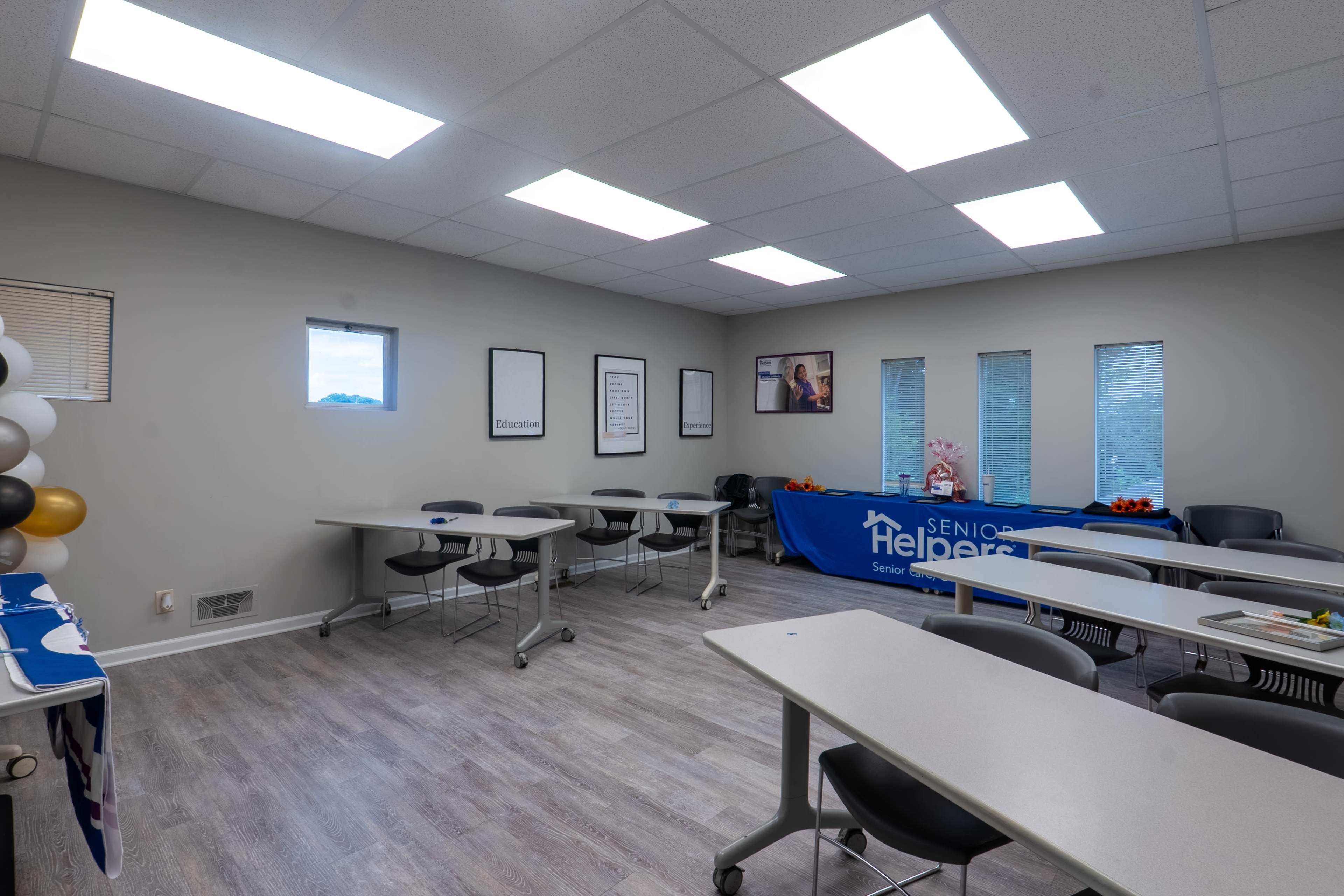 The image depicts a room set up for a meeting or seminar, featuring several tables arranged in rows, a backdrop with a logo, and large windows providing natural light.