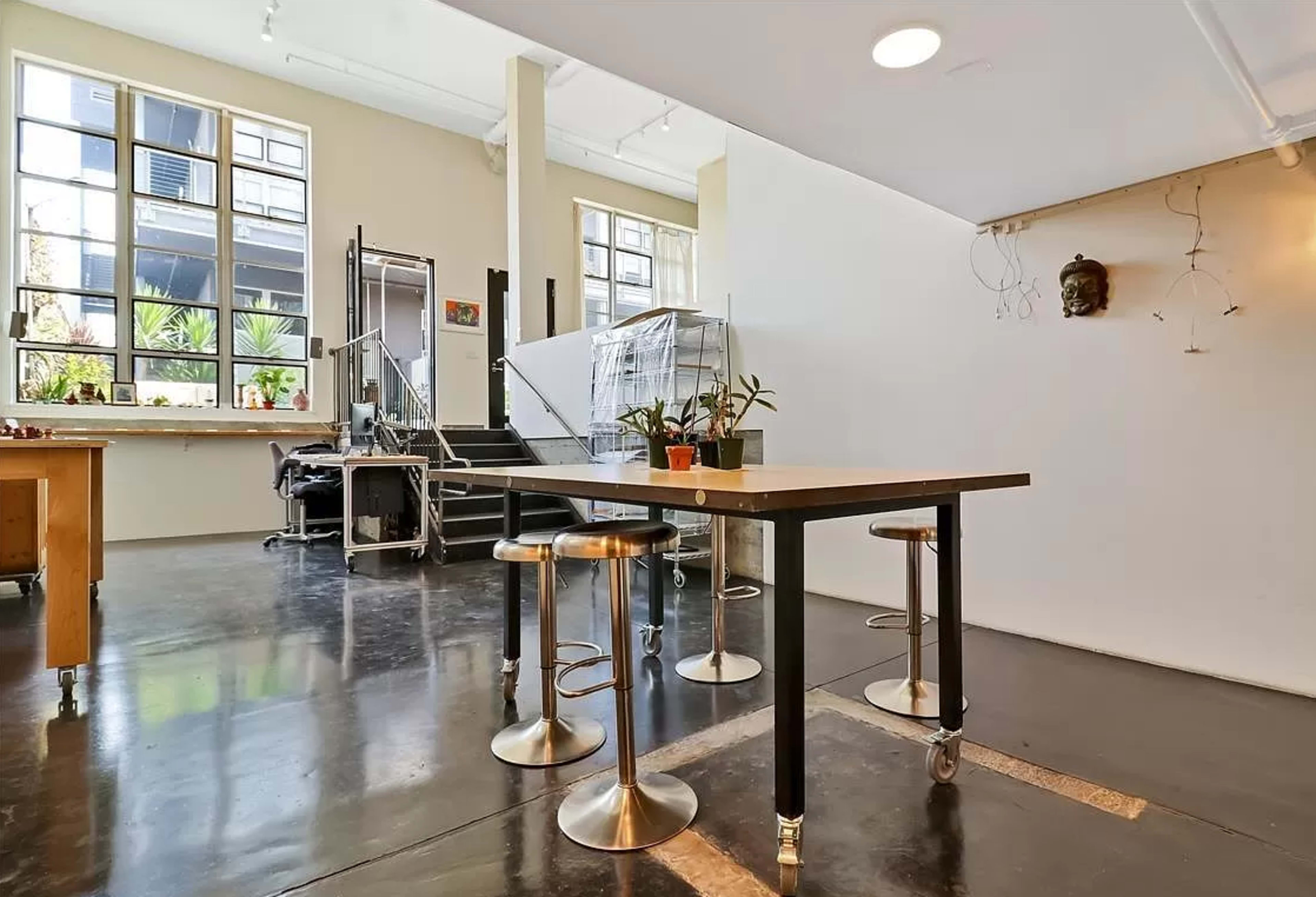 A modern interior space featuring an open layout with a table and stools, a large window letting in natural light, and a staircase in the background.
