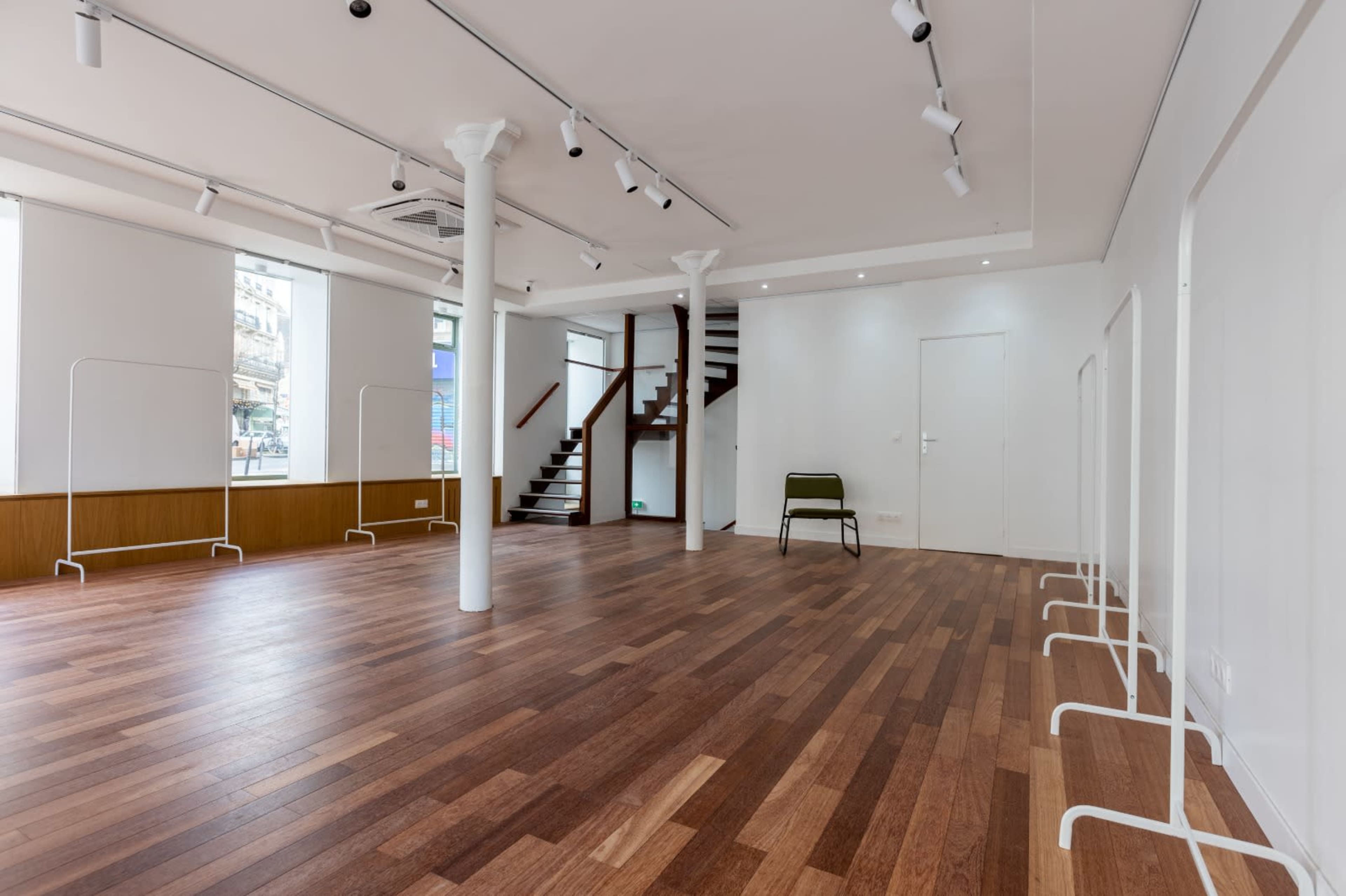 The image shows an empty interior space with wooden flooring, white walls, and a staircase in the background, featuring minimal furnishings and several light fixtures.