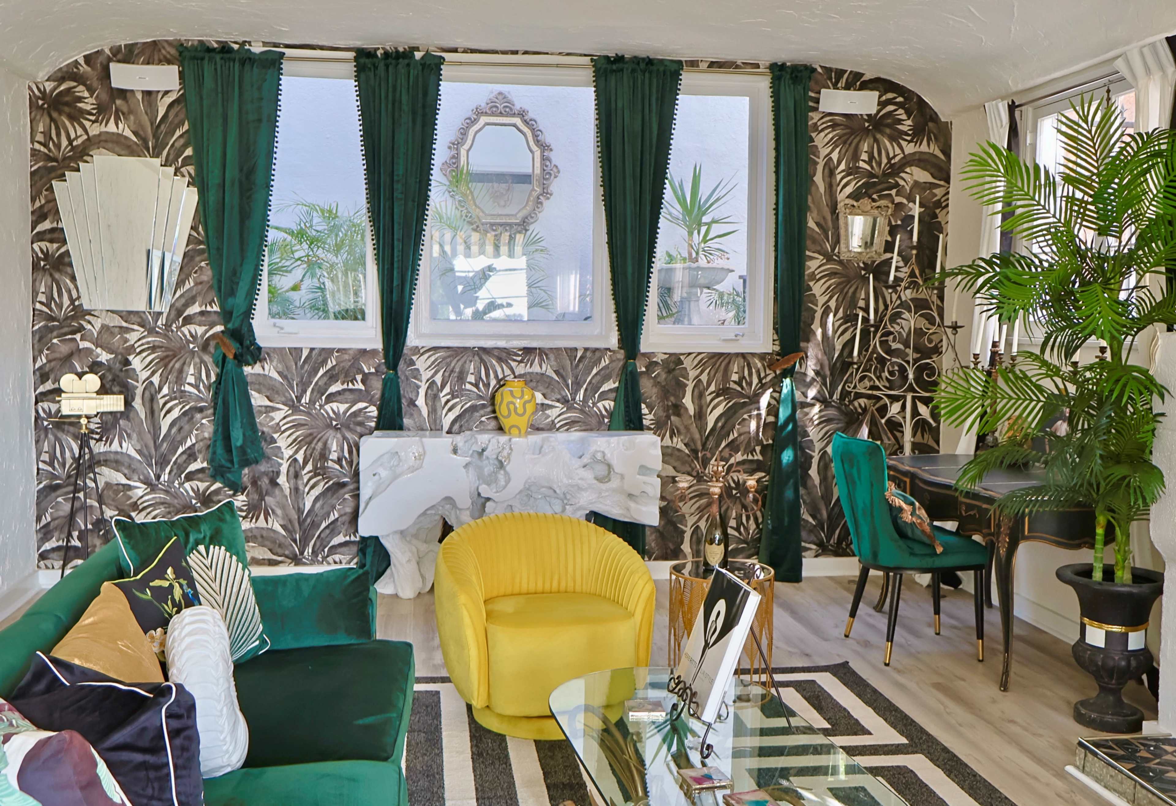 The image shows a stylish living room featuring green curtains, a yellow armchair, tropical-themed wallpaper, and decorative plants.