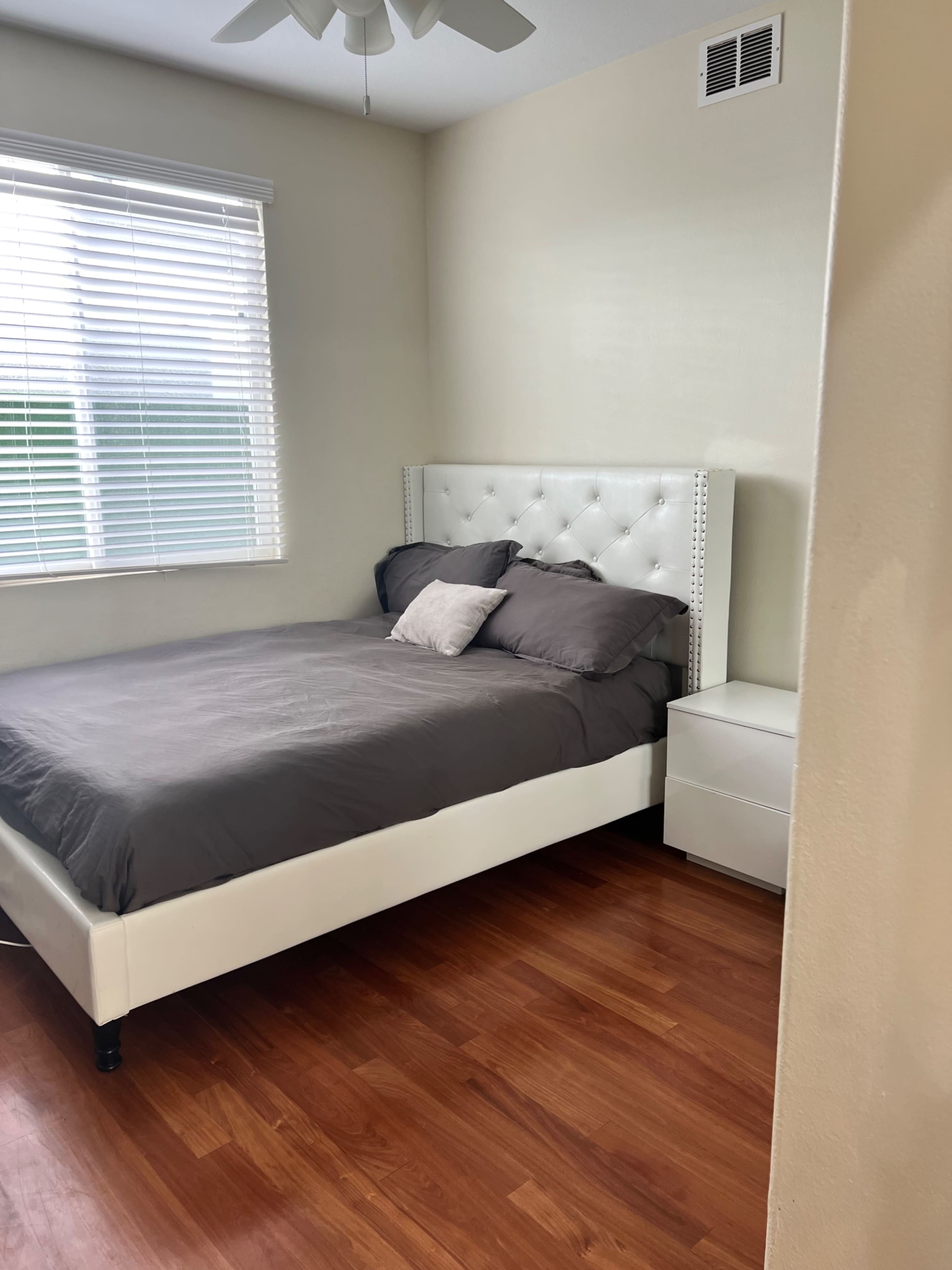 A simple bedroom with a white upholstered bed, gray bedding, a nightstand, and a window with blinds.