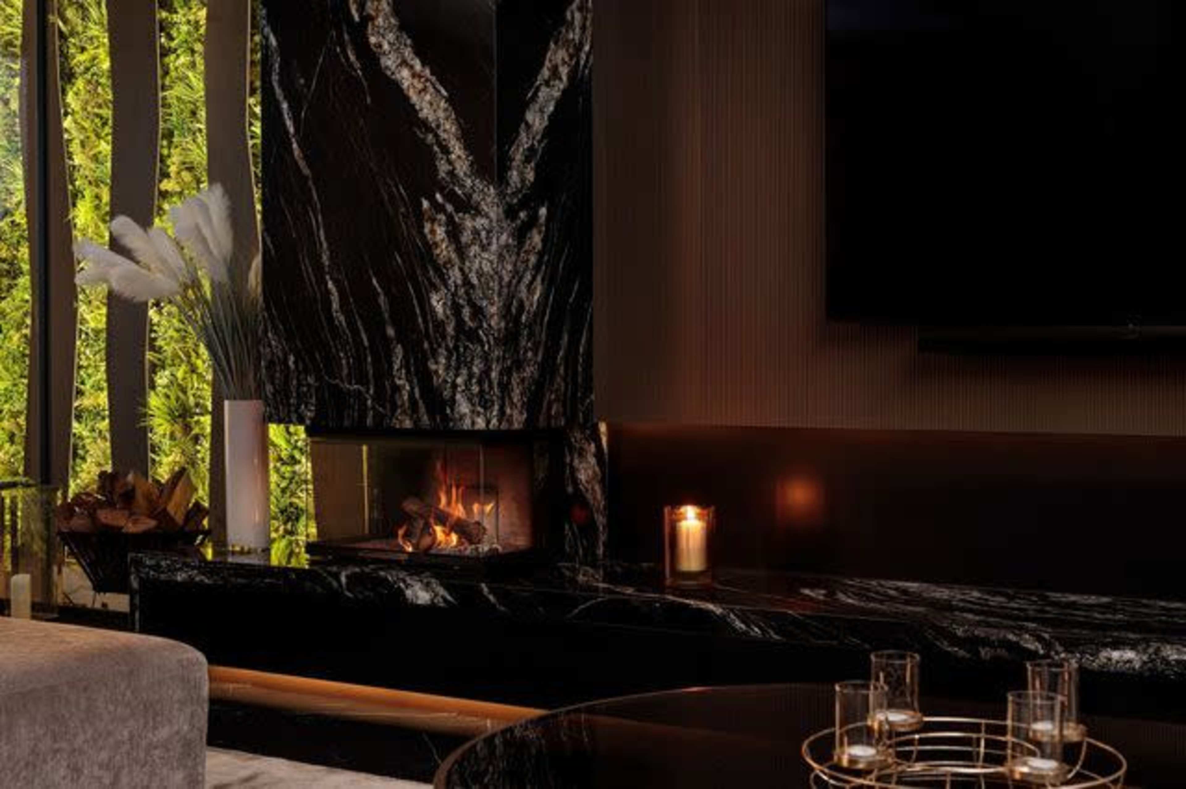 A modern living room featuring a sleek black marble fireplace with a lit candle next to it, surrounded by green plants and contemporary furniture.