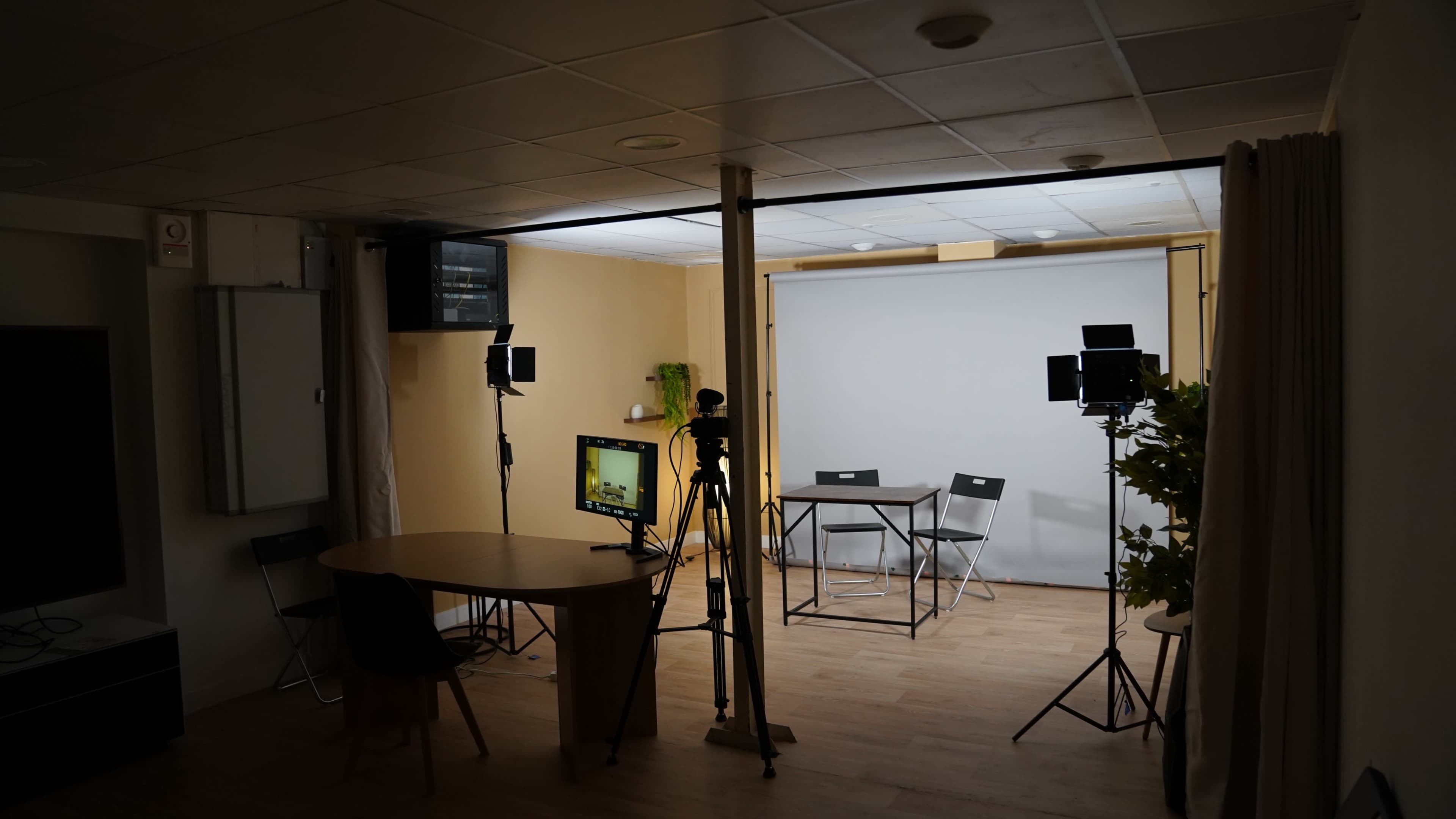 The image shows a small studio setup with a blank backdrop, a table with chairs, and several lights and cameras positioned around the space.