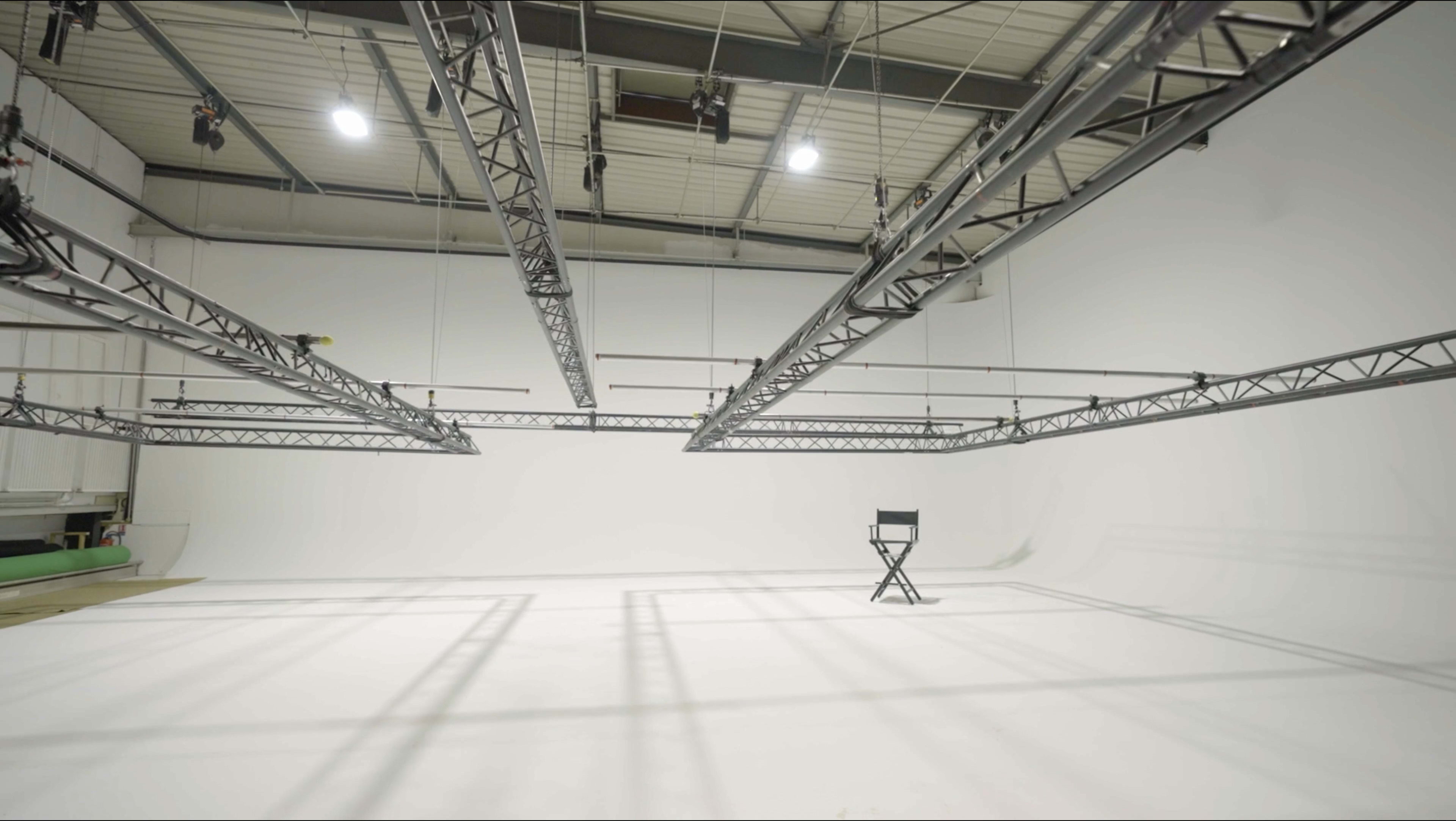 The image shows a spacious film studio with a white backdrop, suspended lighting equipment, and a single director's chair positioned on the floor.