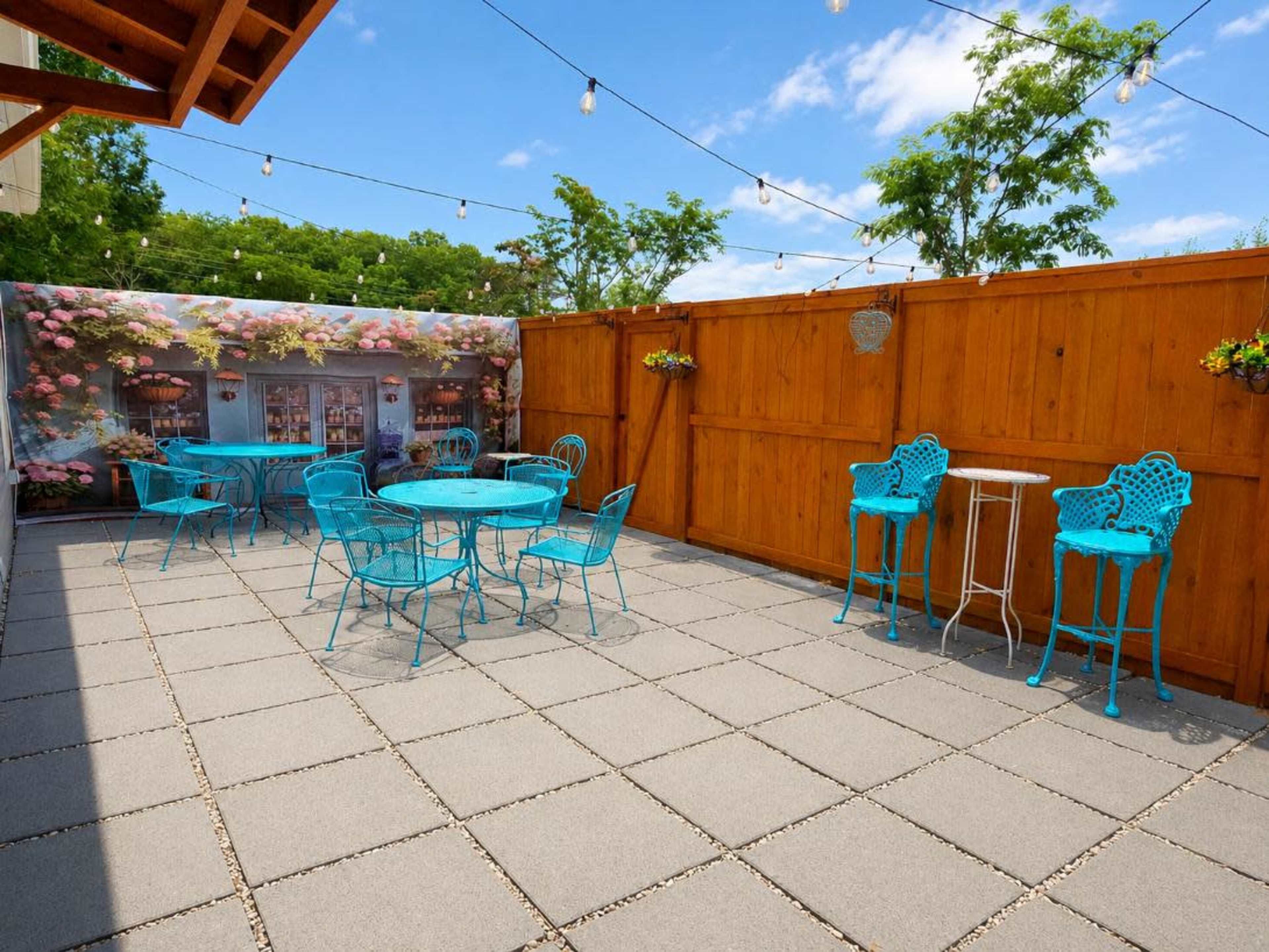 The image shows a patio area with turquoise furniture, including tables and chairs, set against a wooden fence and adorned with string lights and potted plants.