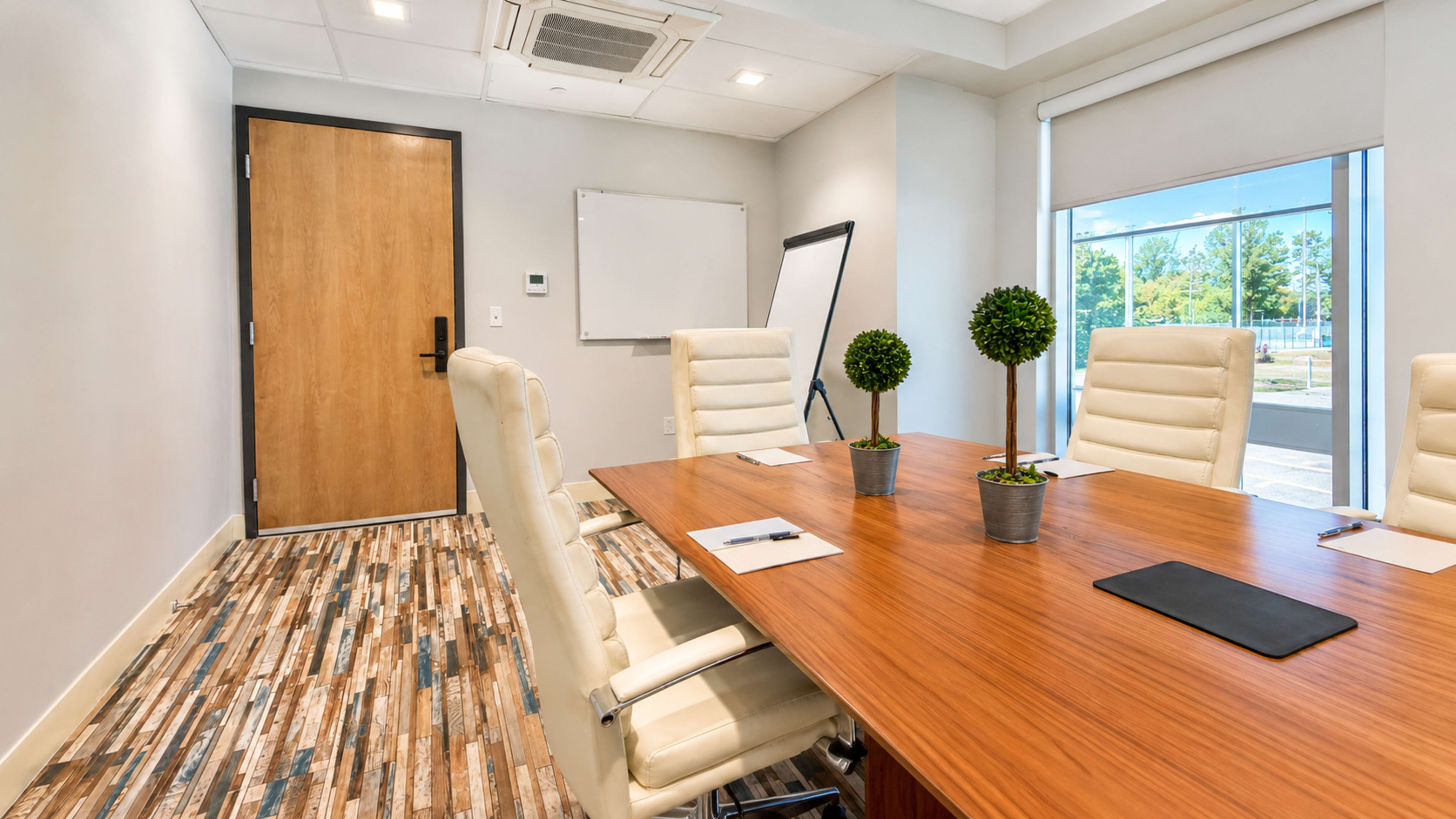 The image shows a modern conference room with a large wooden table, five white leather chairs, two potted plants, and a whiteboard against a gray wall.