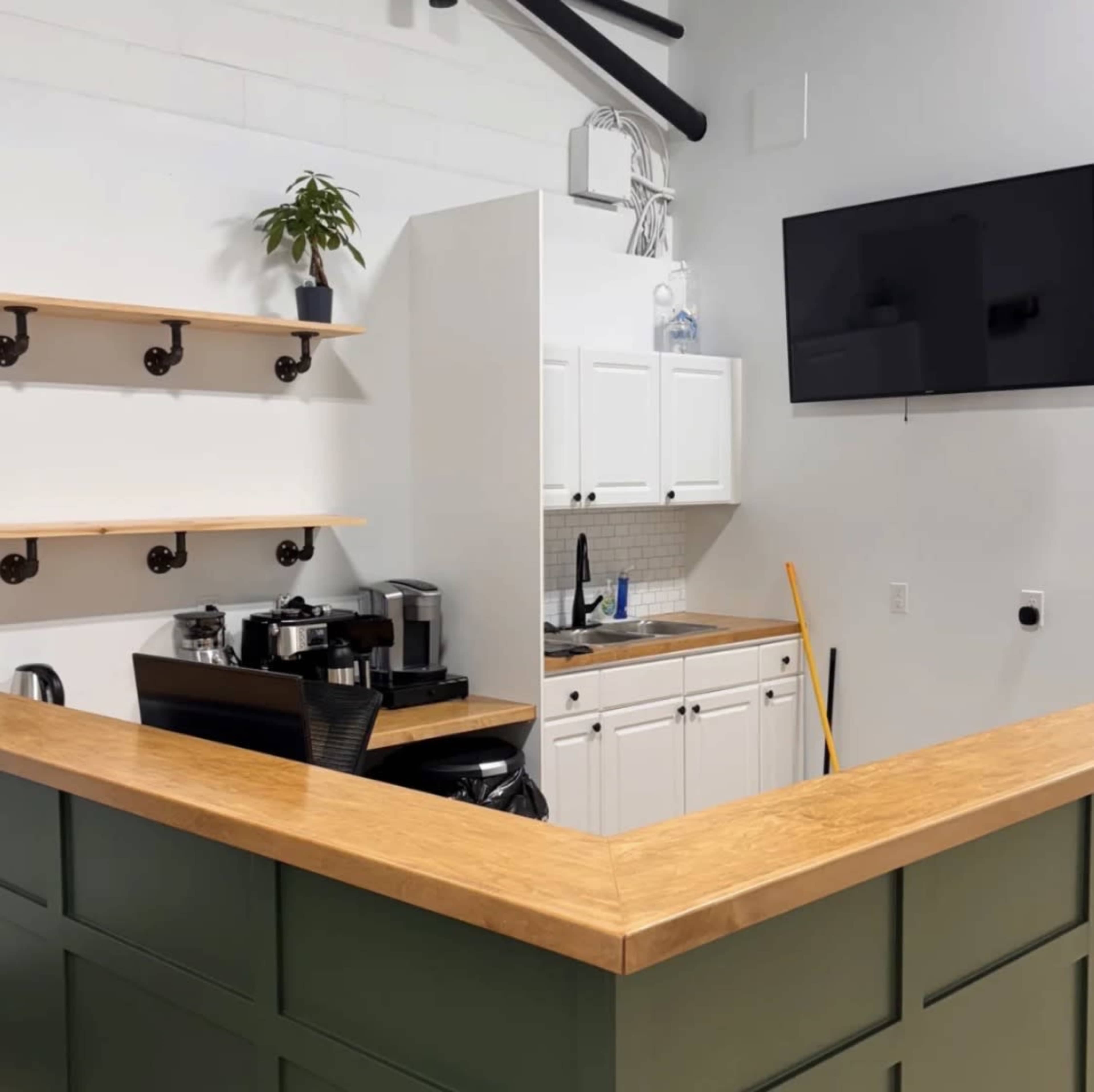 The image shows a small kitchen area with a countertop, shelving, and appliances, including a coffee maker and a sink, within a modern space.
