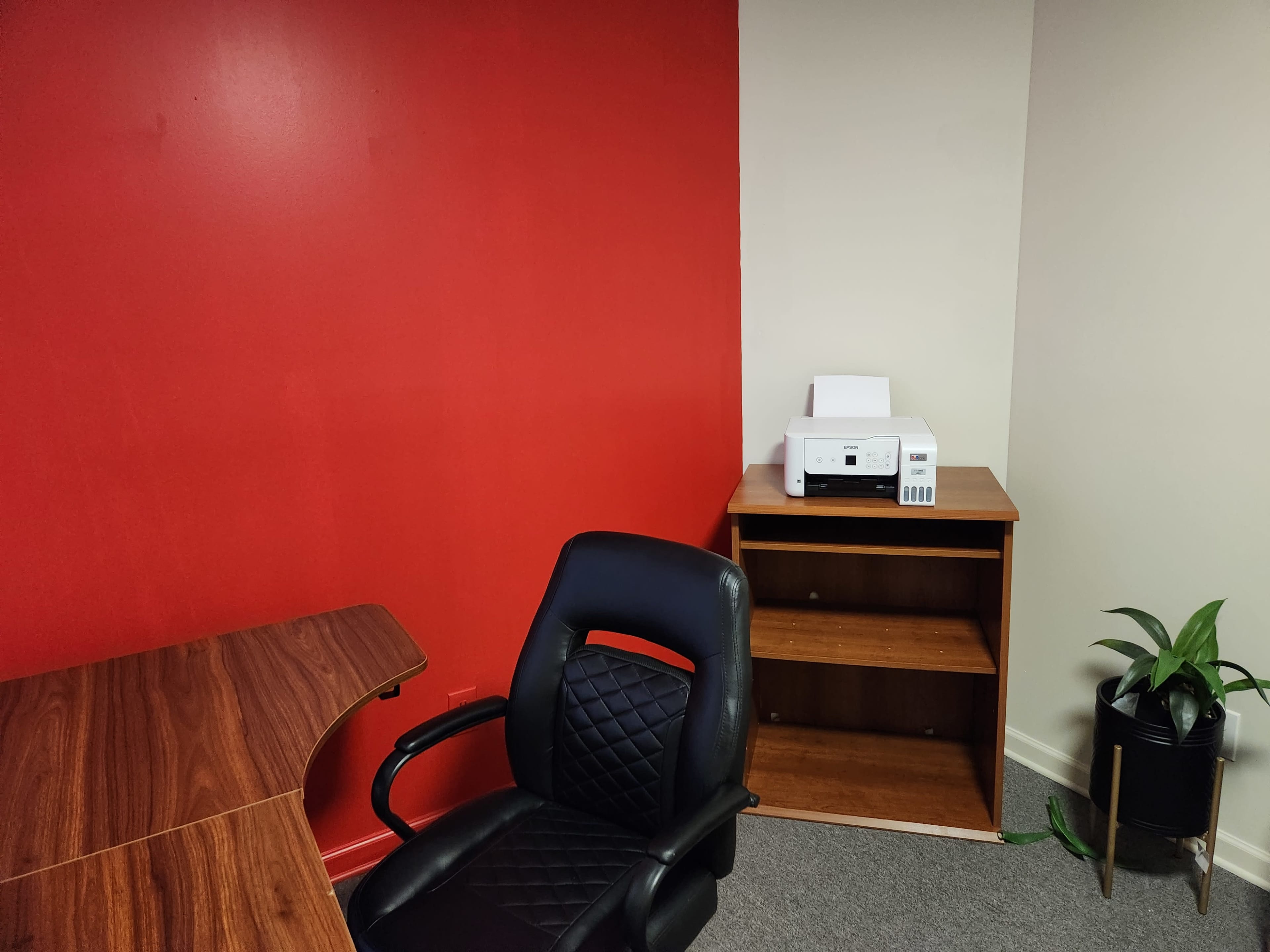 An office space with a red wall, a wooden desk, a black chair, a printer on a wooden shelf, and a potted plant in the corner.