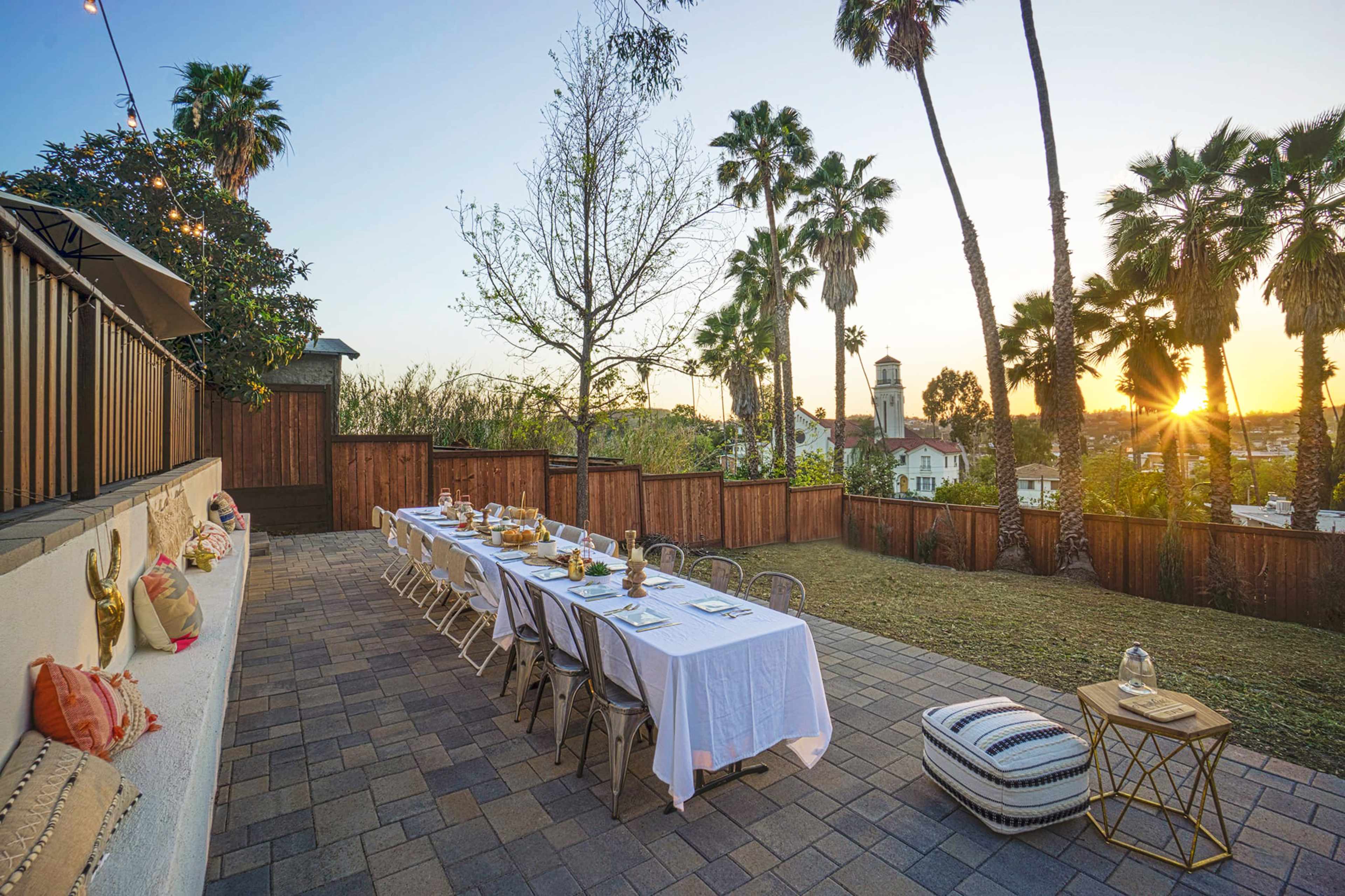 A long outdoor dining table is set with white linens and dinnerware, surrounded by palm trees at sunset.