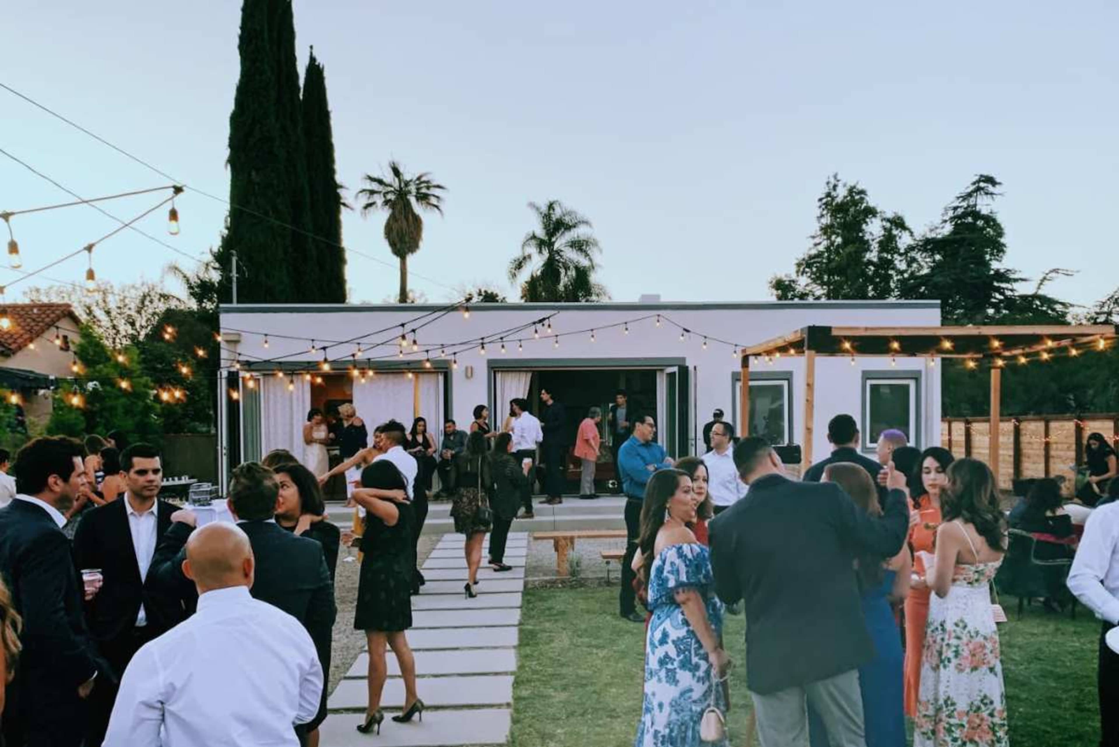 A gathering of people in formal attire mingles in a backyard setting with string lights and a modern house in the background.