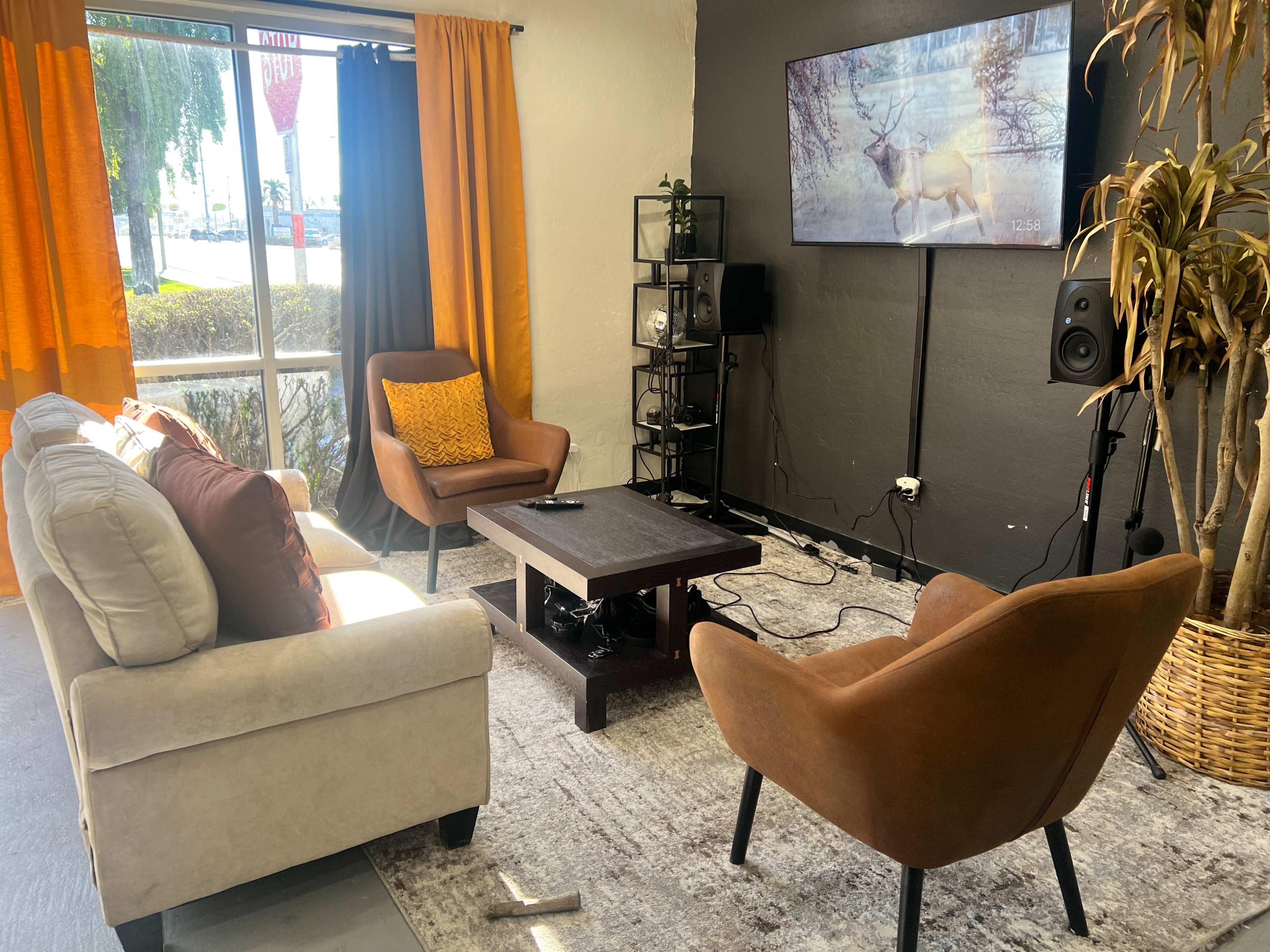 A cozy living room features two armchairs, a sofa, a coffee table, and a television mounted on the wall, with large windows letting in natural light.