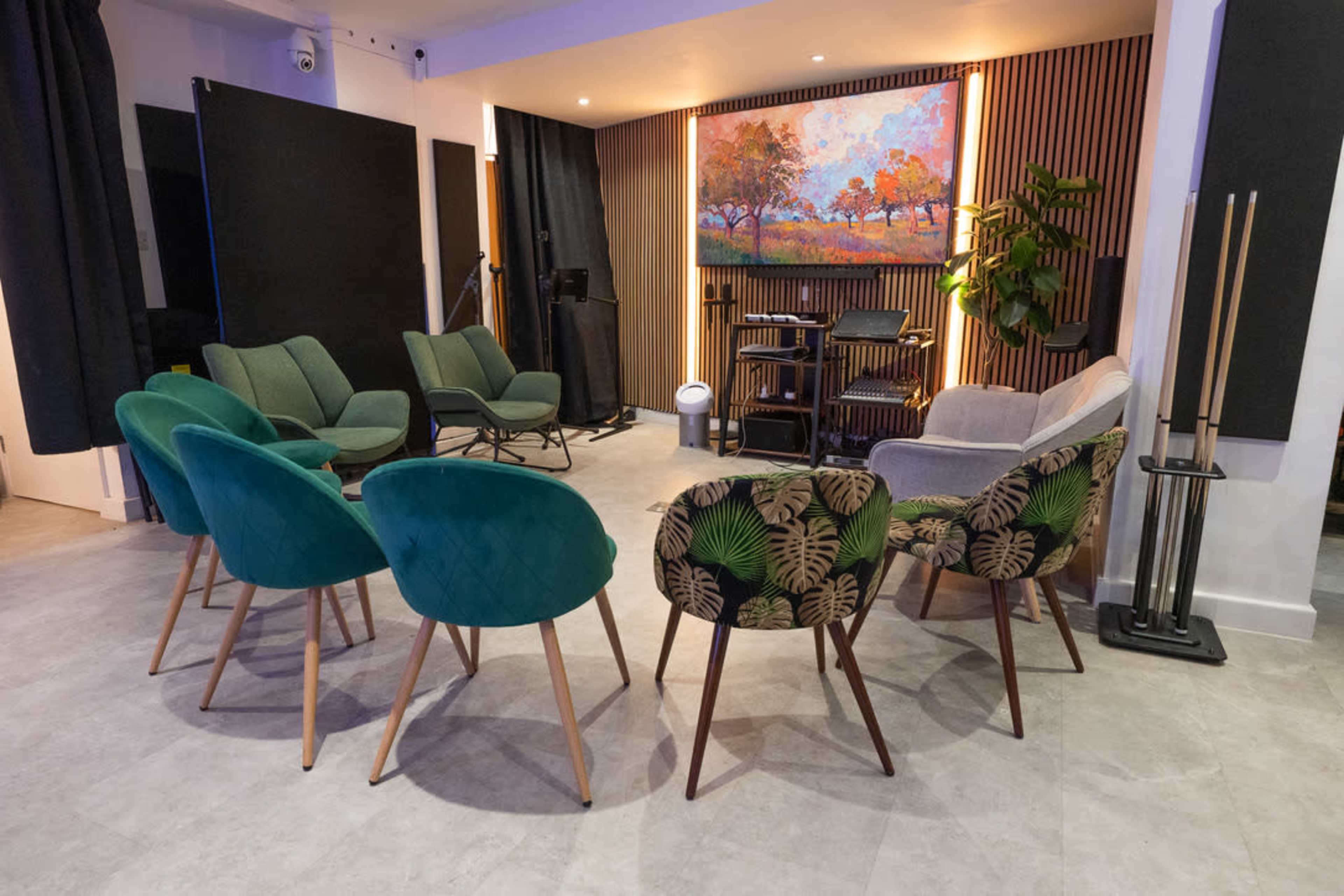 A cozy seating area features green and patterned chairs arranged in a circle, with a colorful painting displayed on the wall behind them.