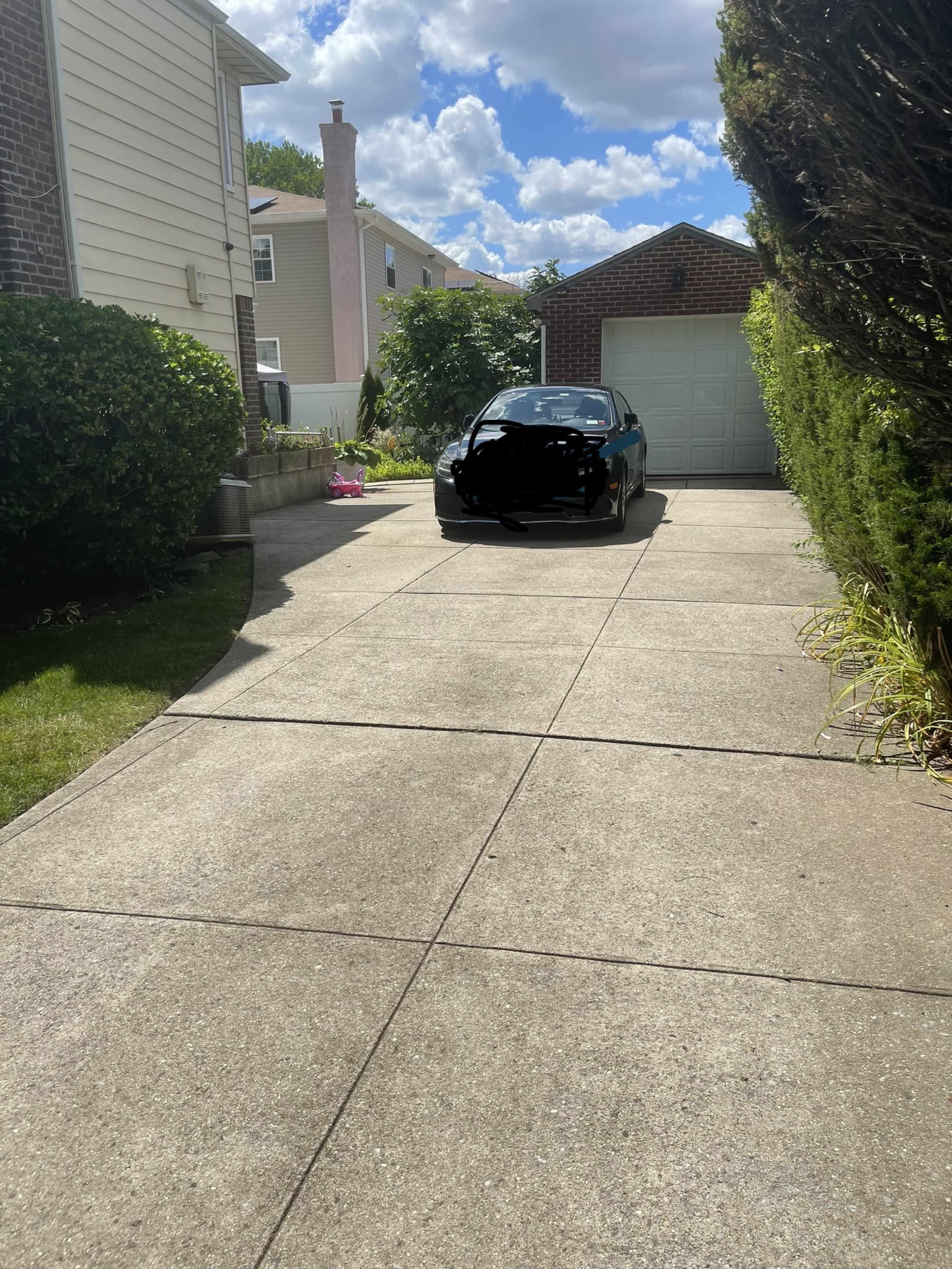 A black car is parked in a concrete driveway leading to a garage, bordered by shrubs and houses on either side.