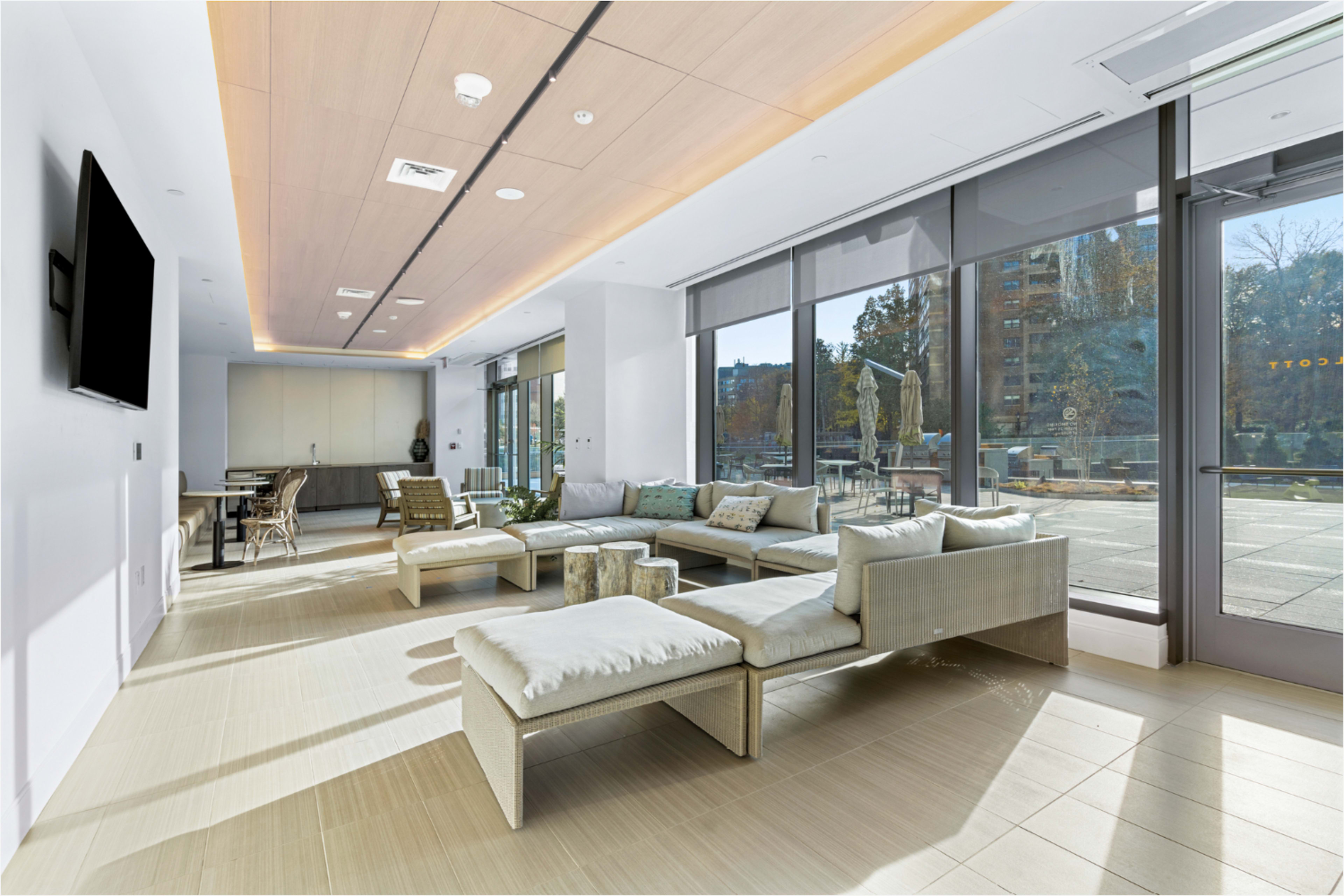 The image shows a modern lounge area with a sectional sofa, coffee table, and large windows that let in natural light.