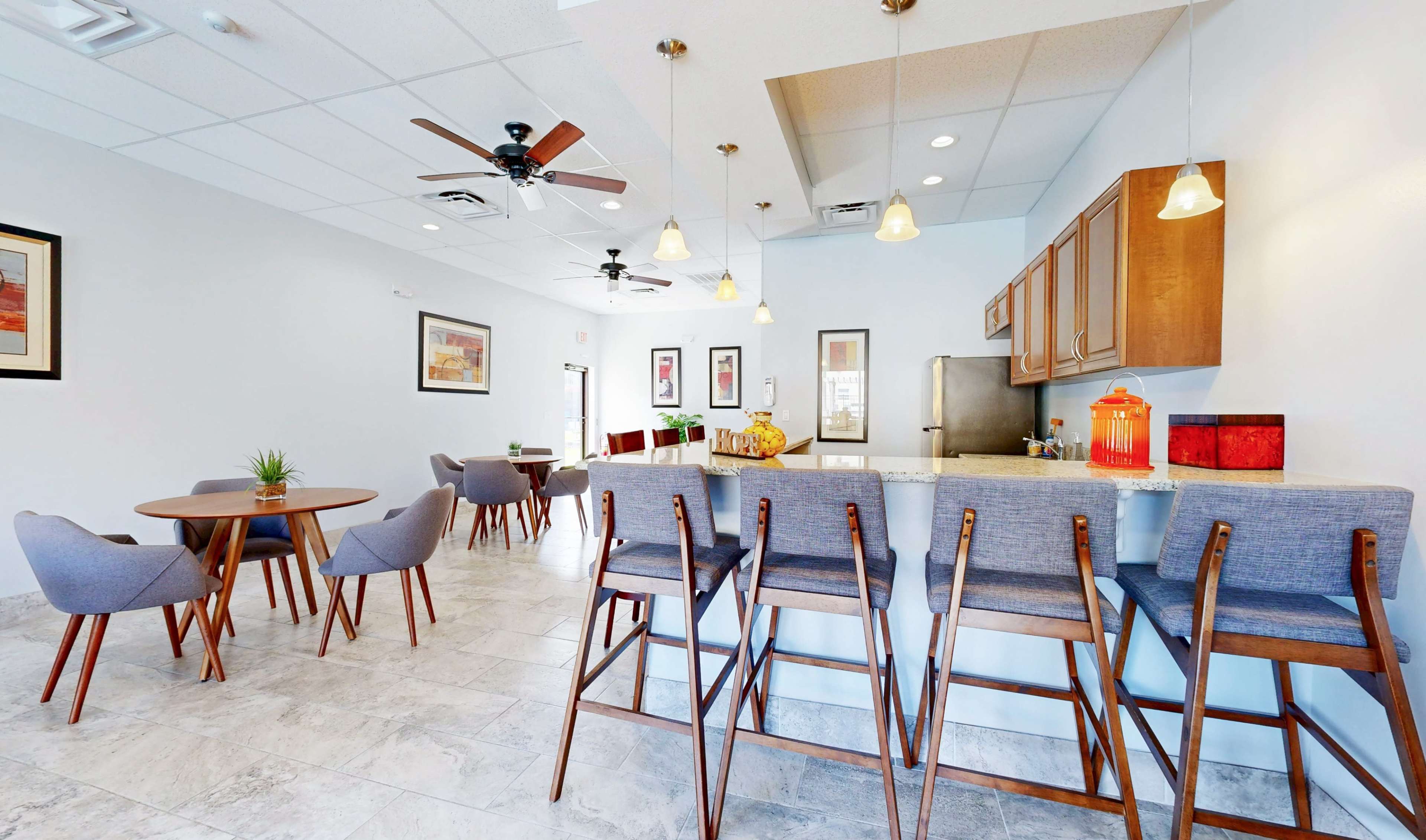 The image shows a light-filled room with modern furniture, including a bar area with high stools, a dining table, and a kitchenette.