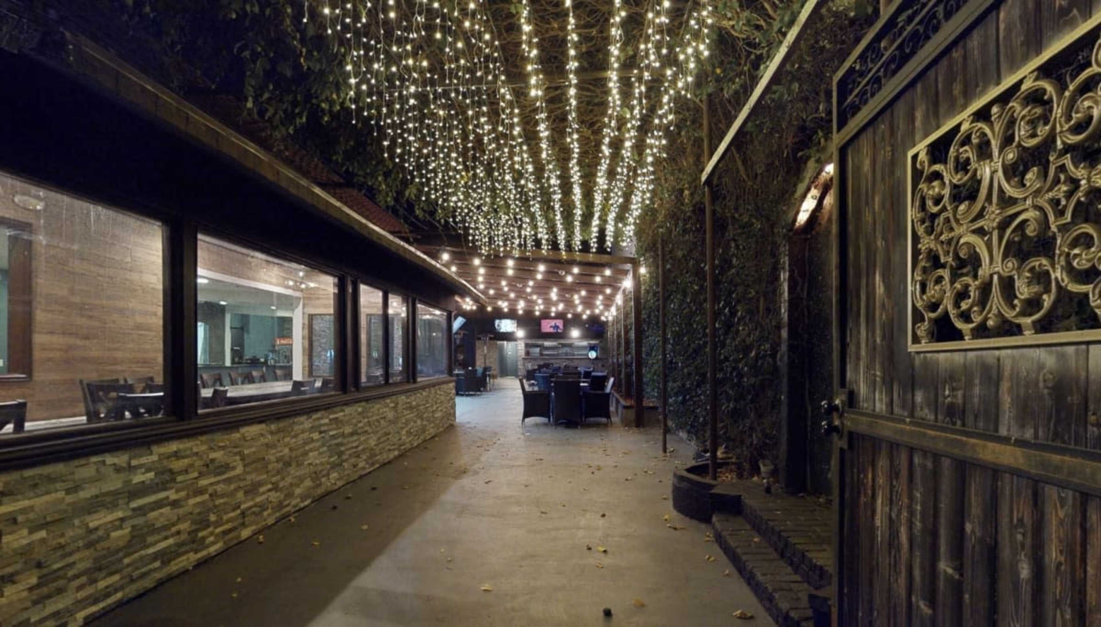 The image shows a pathway leading to a dining area, illuminated by strings of lights overhead and bordered by greenery.