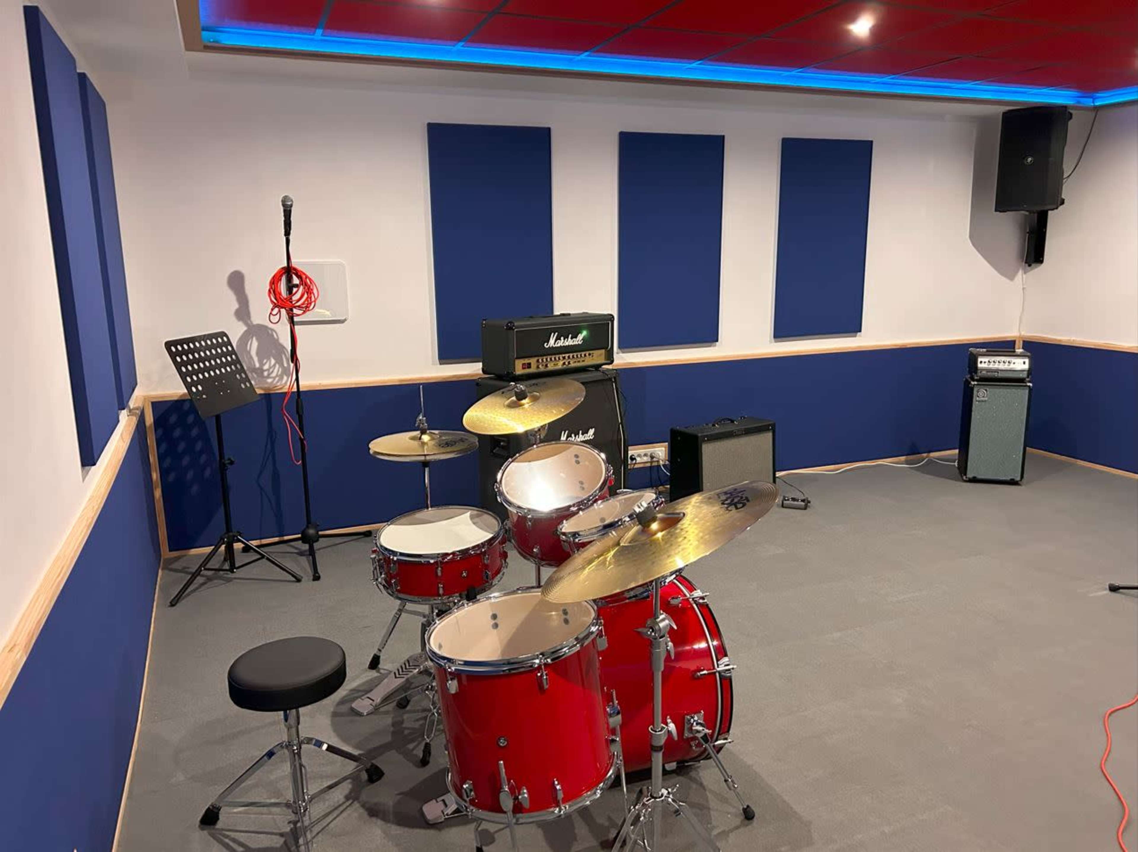 A drum set in a rehearsal space is positioned next to amplifiers and a microphone stand, with blue soundproof panels on the walls.