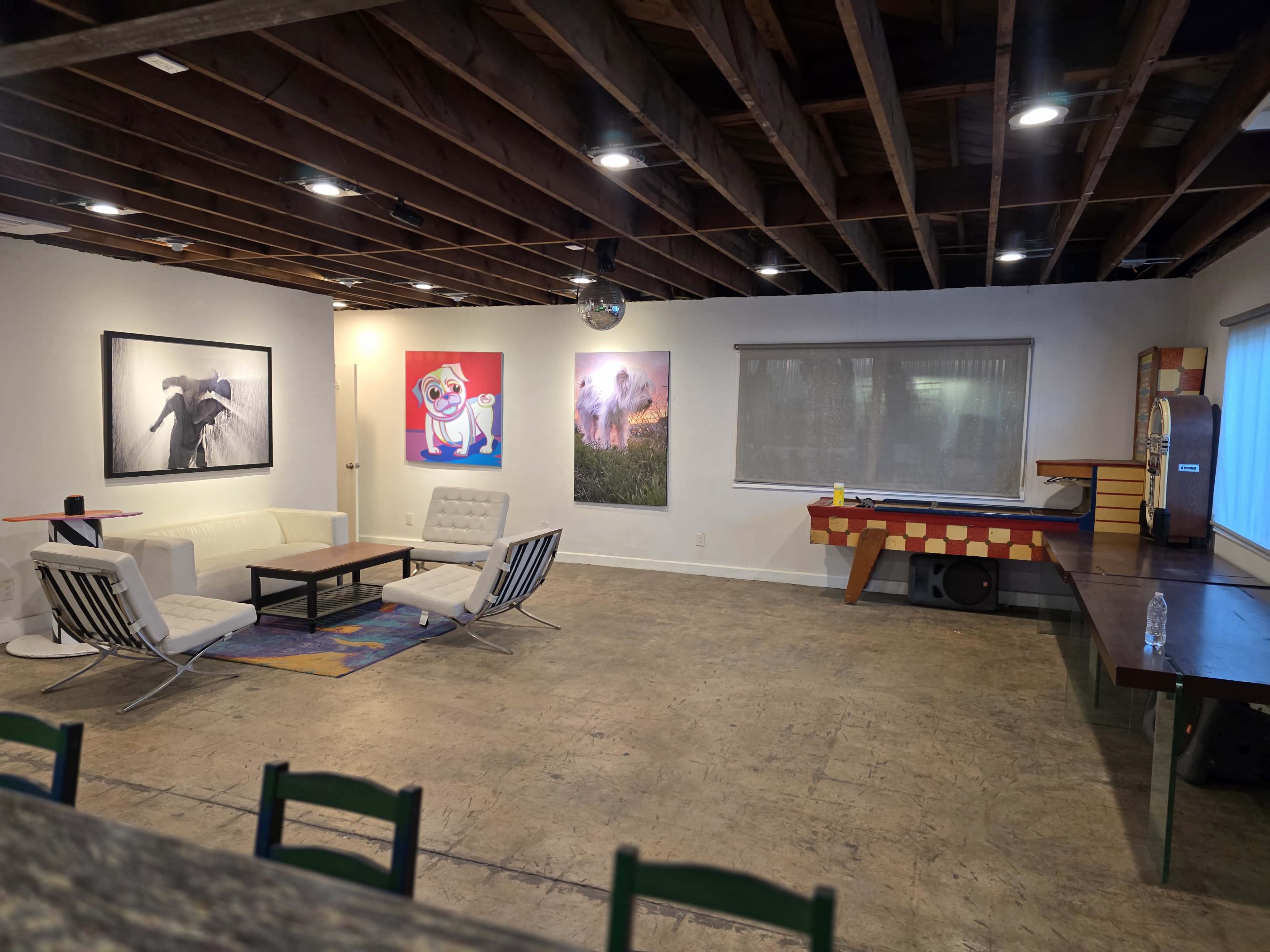 The image shows a spacious room with concrete flooring, featuring a seating area with white sofas, a coffee table, and colorful artwork on the walls.