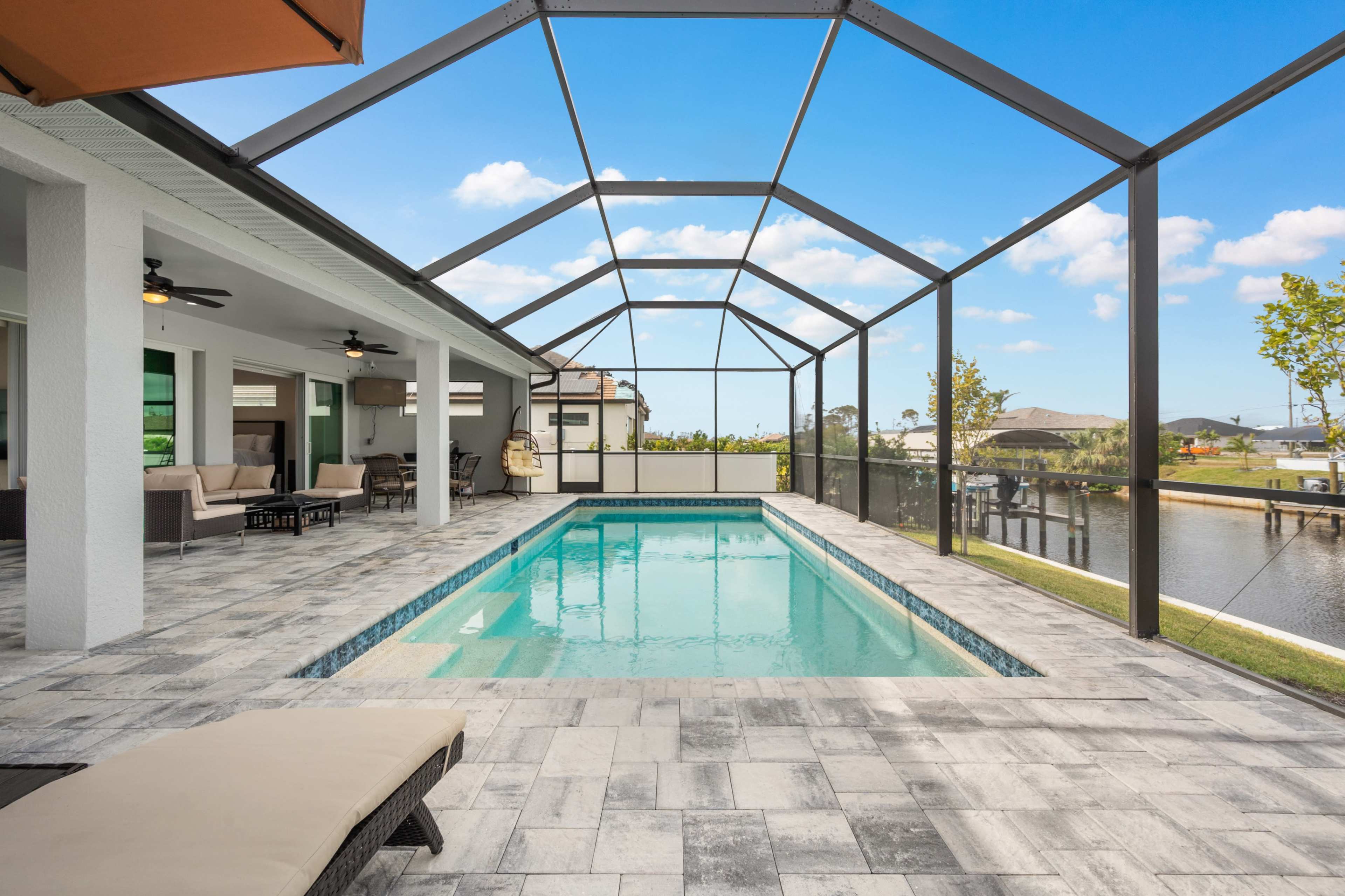 The image shows a screened-in patio with a swimming pool overlooking a body of water and surrounding palm trees.