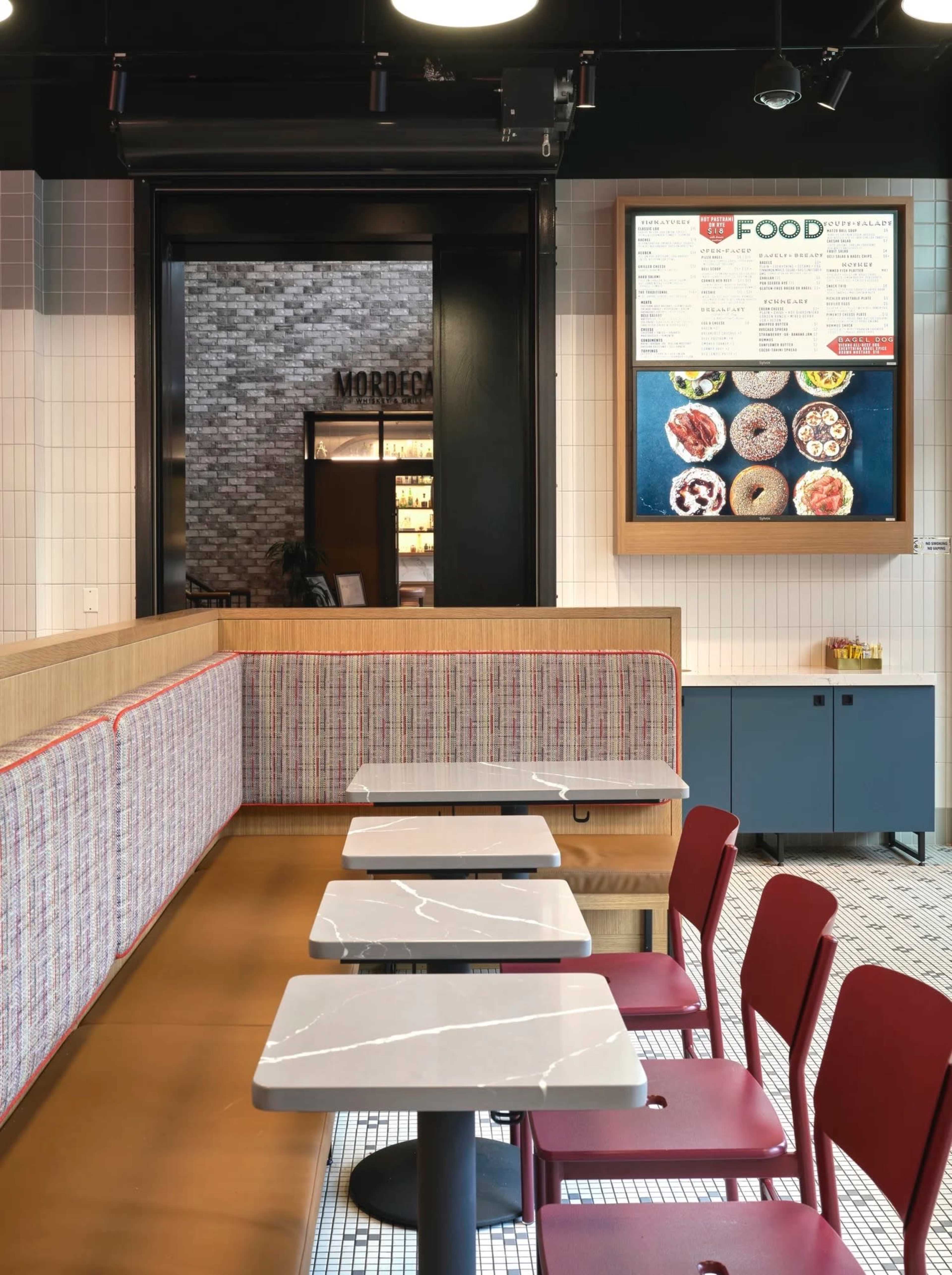 The image shows a modern café interior with empty tables and chairs arranged against a patterned wall, alongside a menu board displaying food options.