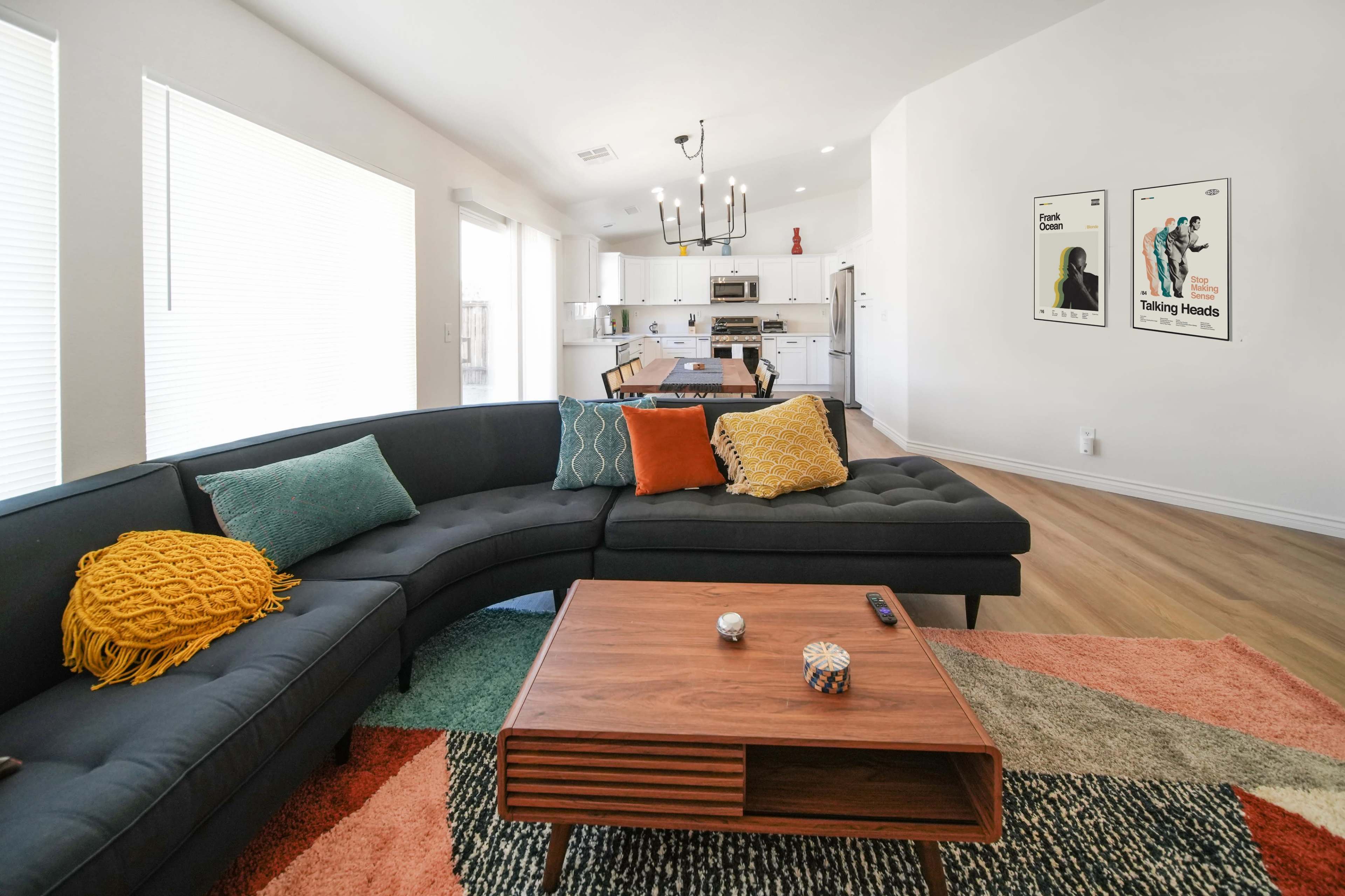 A modern living room features a curved black sectional sofa with colorful cushions, a wooden coffee table, and a dining area visible in the background.