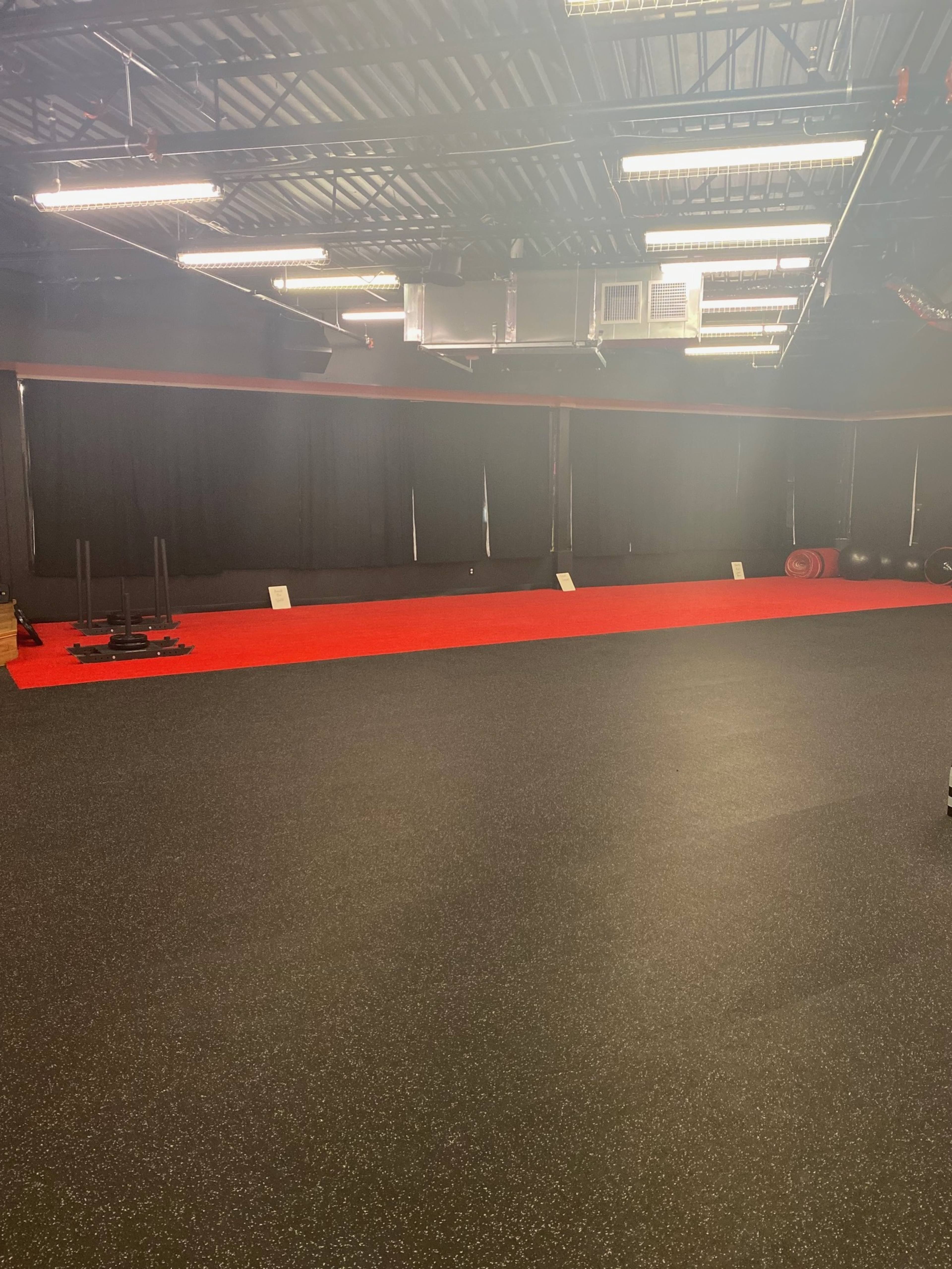 A spacious fitness studio with black rubber flooring, red accents, and black curtains lining the walls.