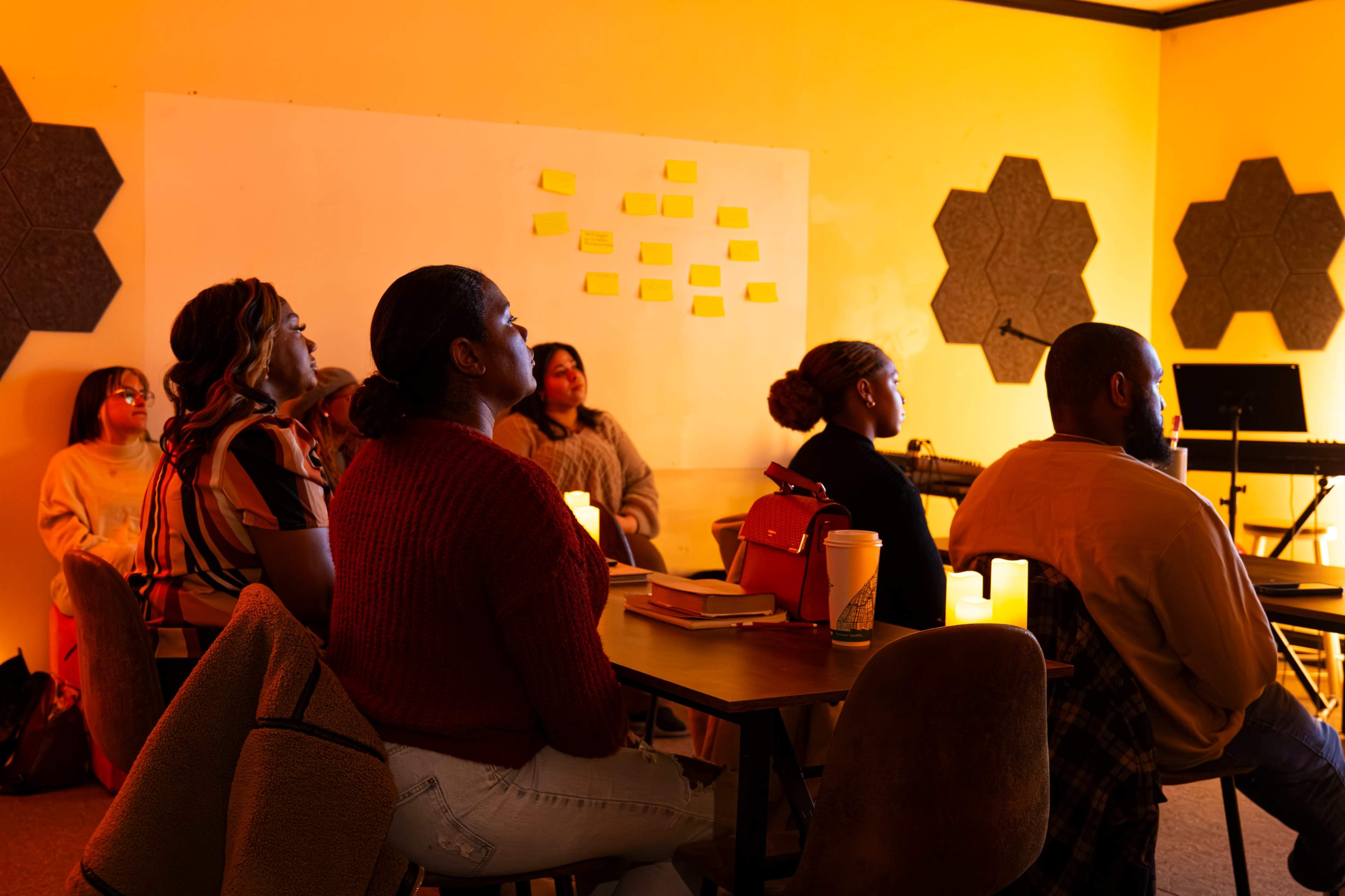 A group of people sits at tables in a warmly lit room, attentively facing a presentation area with a blank wall and sticky notes.