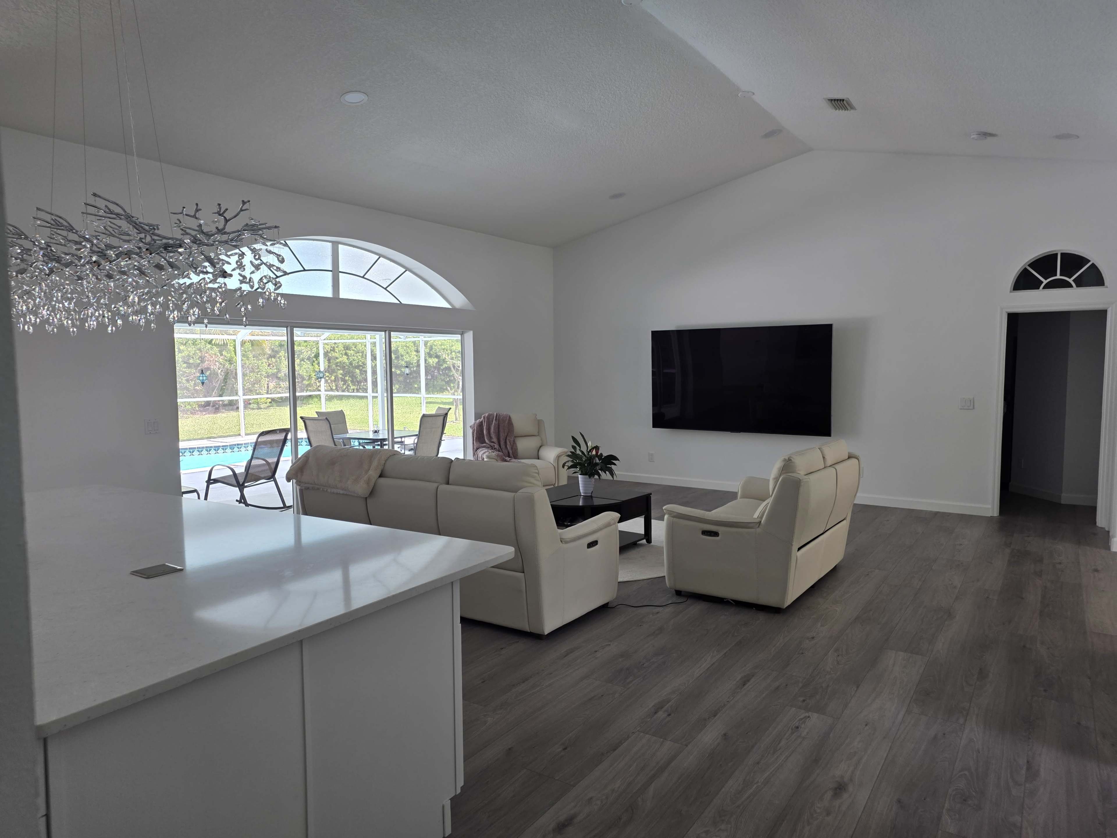 The image shows a modern, open-concept living area with light-colored furniture, large windows letting in natural light, and a view of a pool outside.