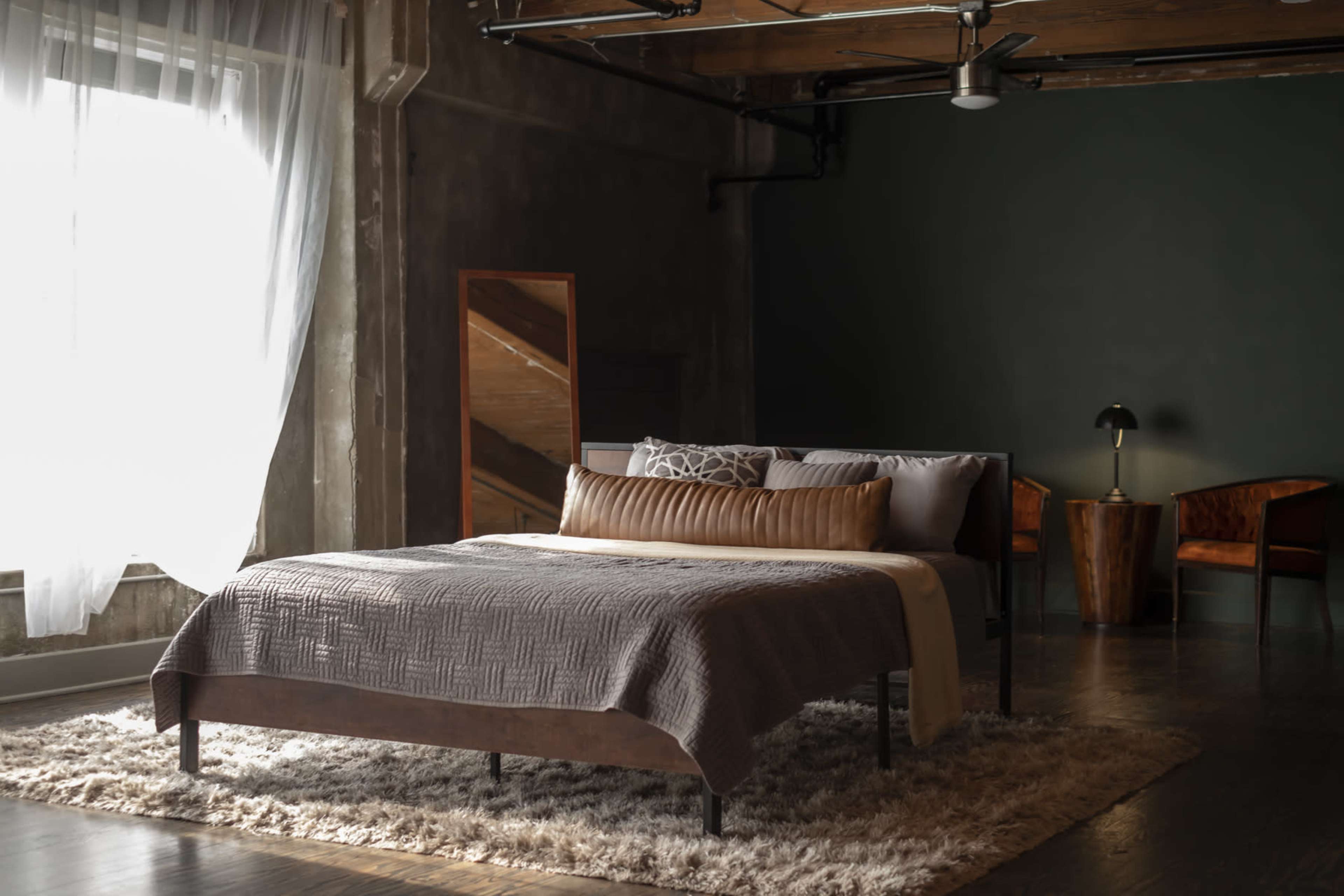 A modern bedroom features a bed with a textured blanket, decorative pillows, and a soft area rug, complemented by large windows and minimalist furniture.