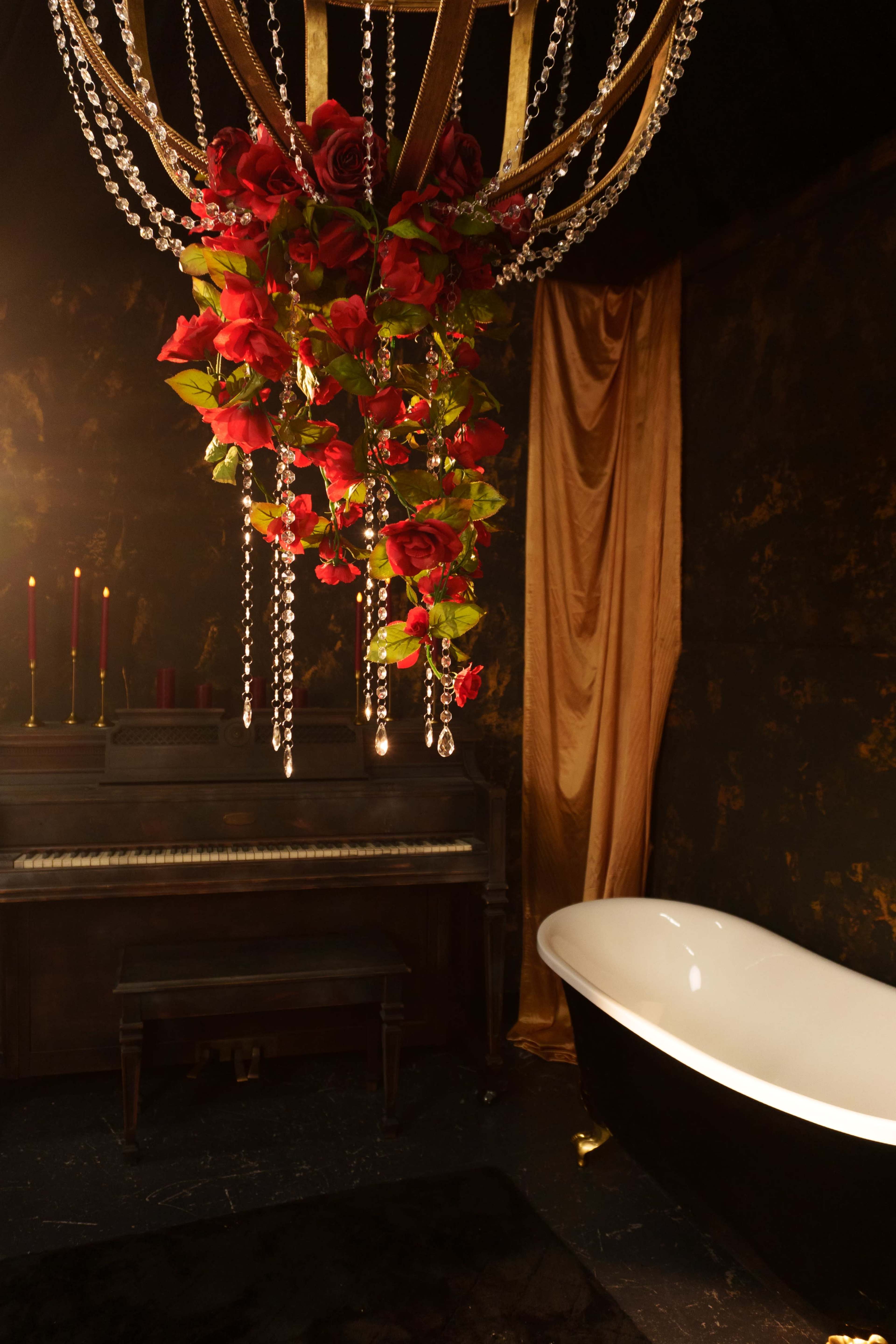 A vintage piano sits next to a black and white clawfoot bathtub, both illuminated by a chandelier adorned with red roses and crystals in a dimly lit room.