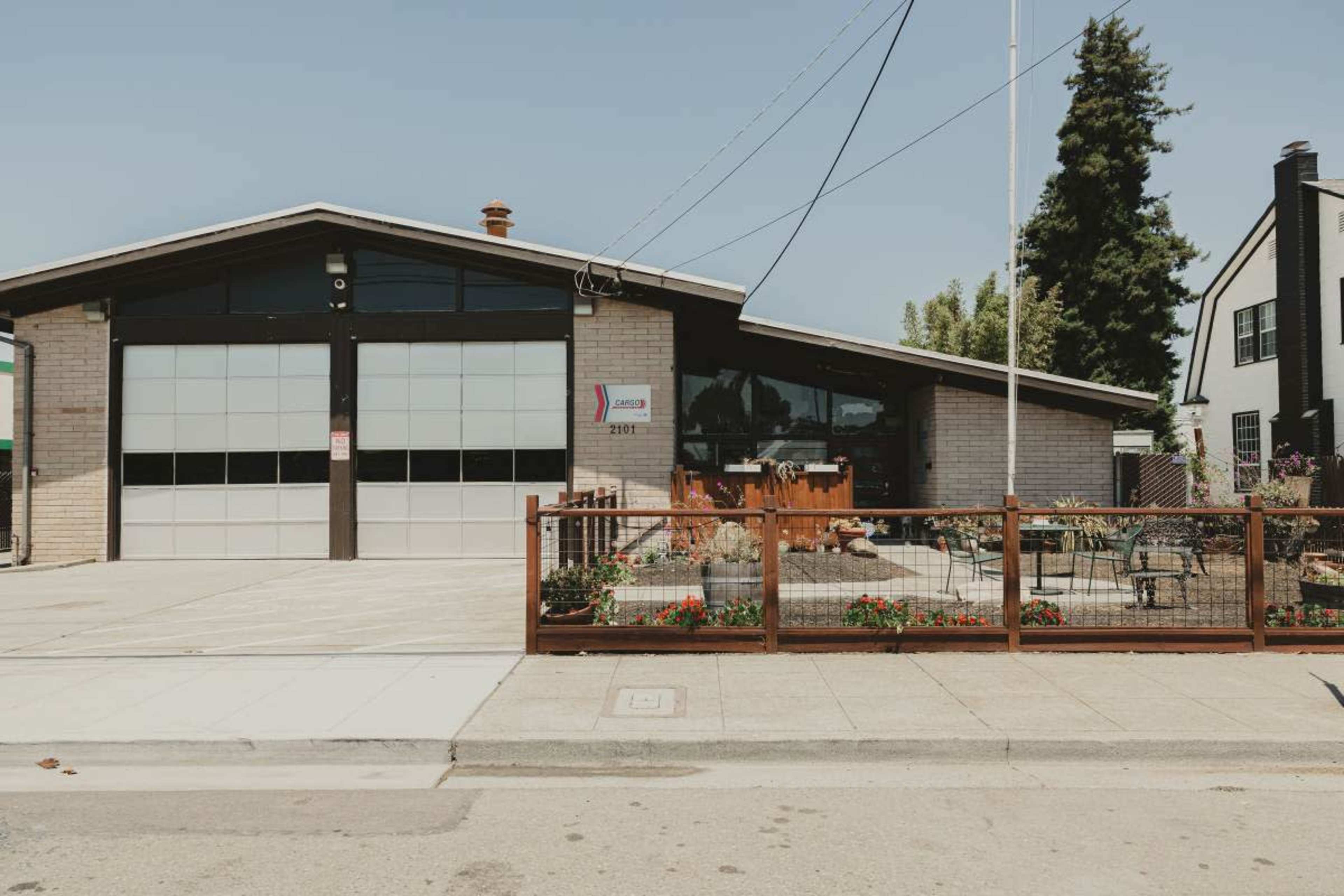A one-story building with large glass garage doors and a landscaped outdoor seating area is located on a paved street.