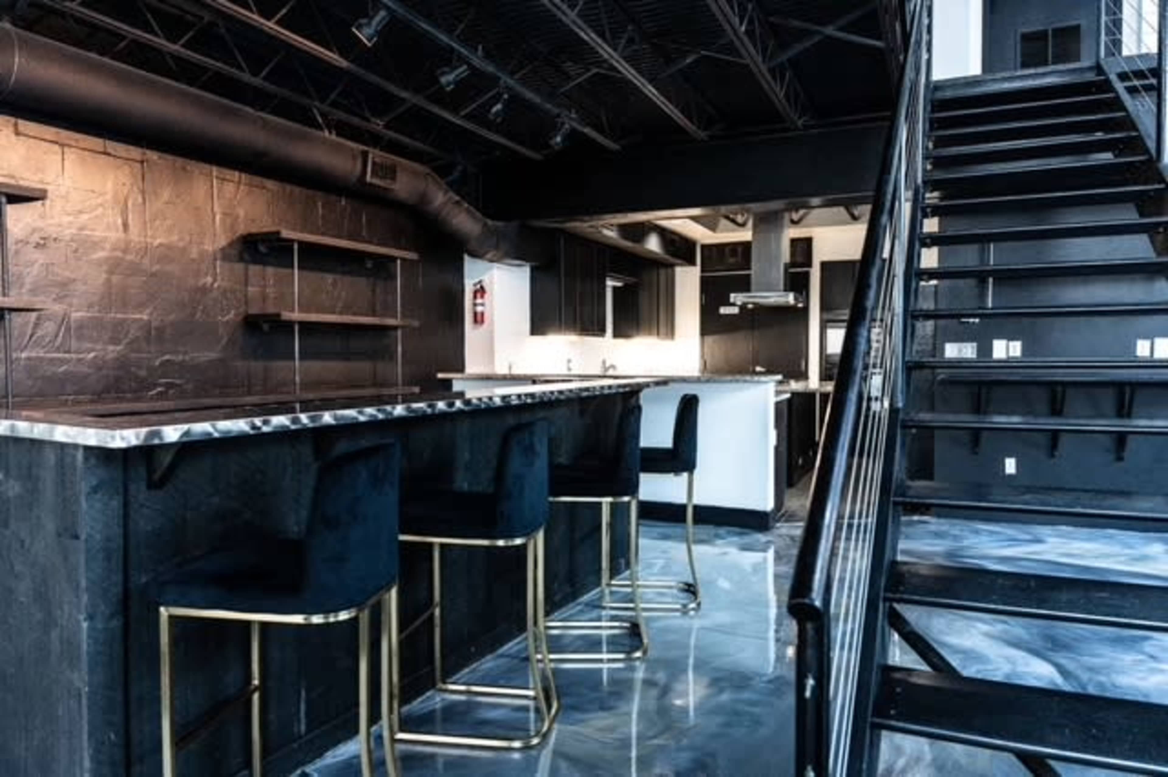 The image shows a modern kitchen and bar area with black cabinetry, metallic accents, and a staircase leading to an upper level.