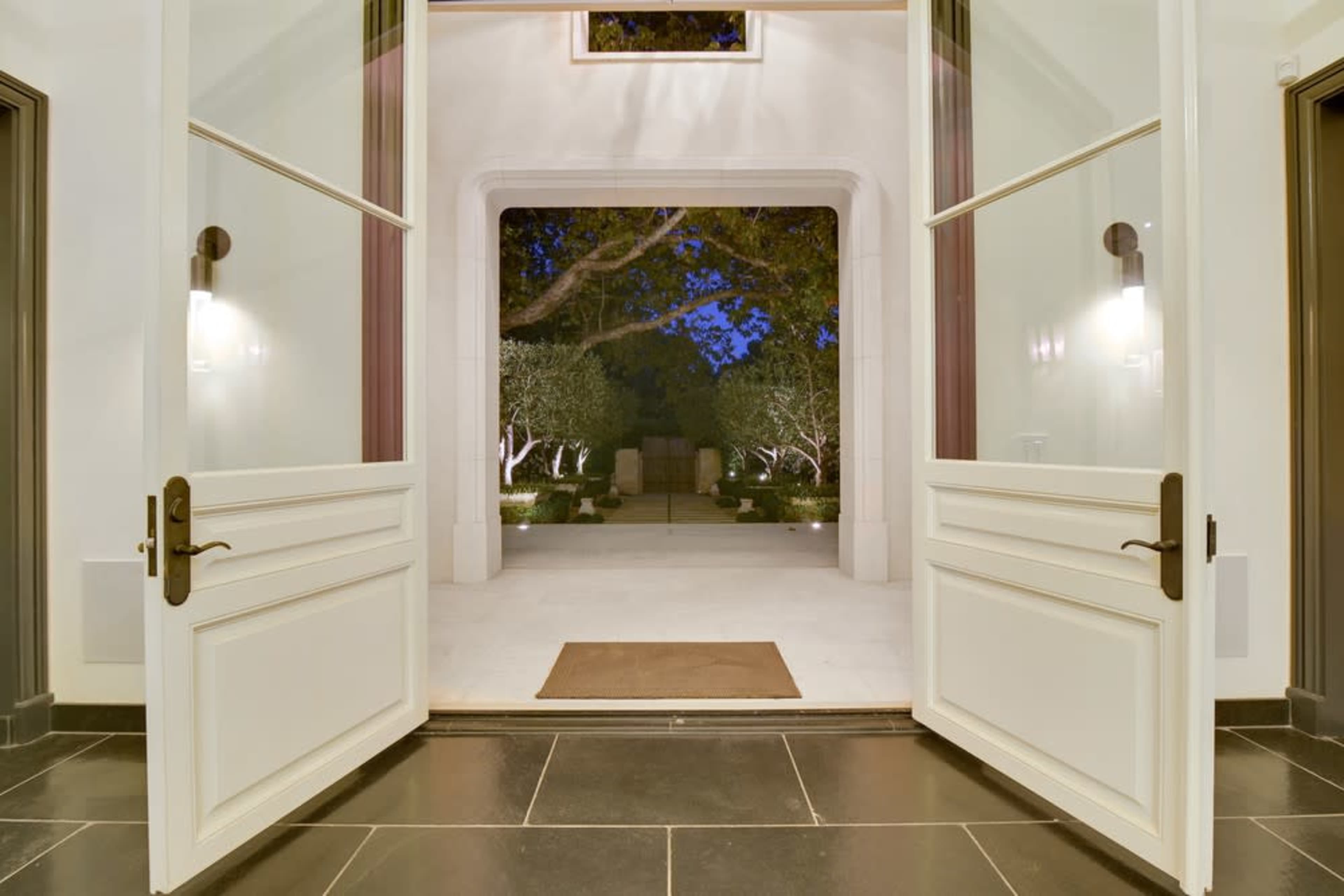 An entrance with large double doors open, revealing a well-lit outdoor space with trees and stone features in the background.
