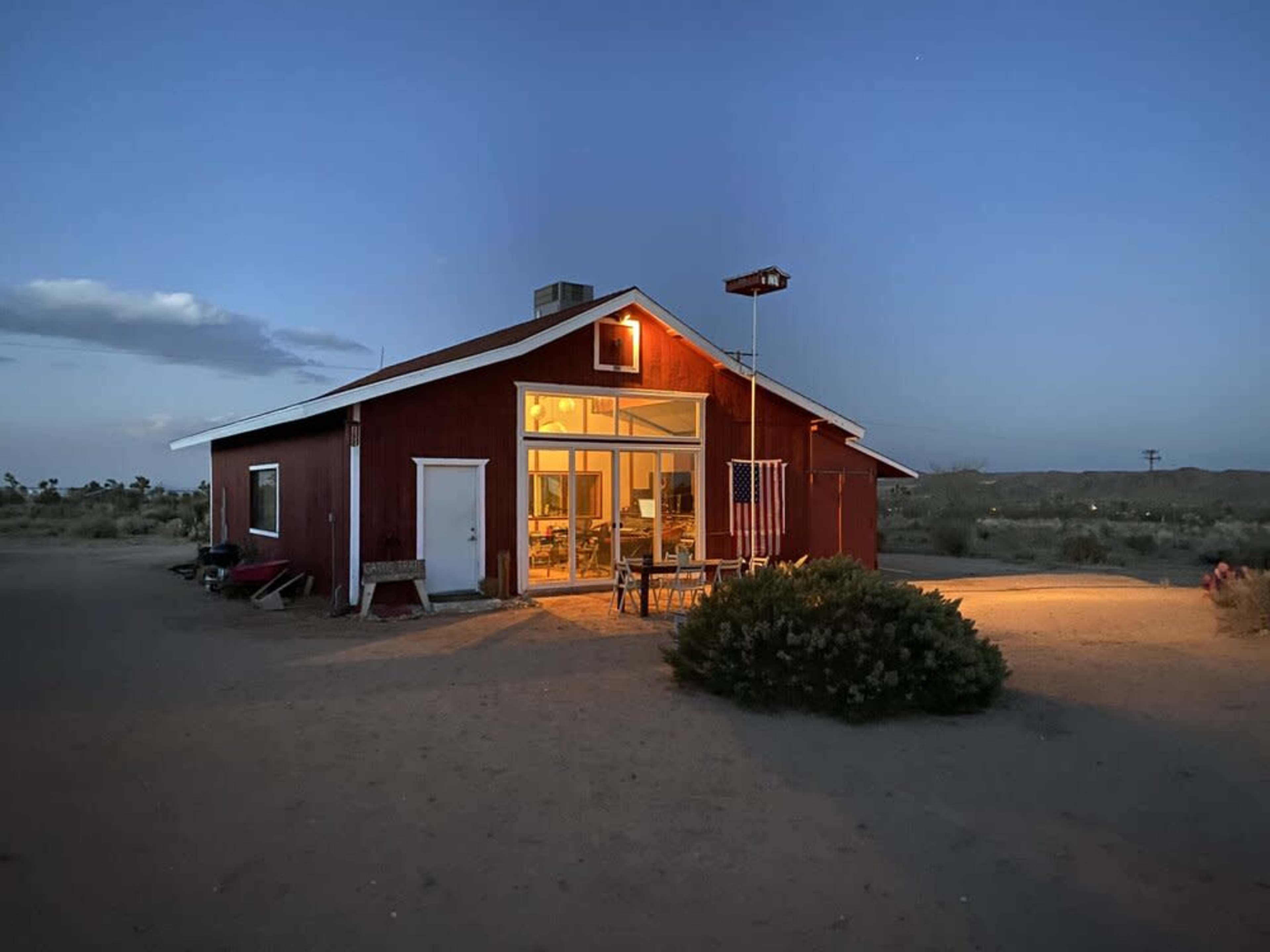 A red farmhouse with large windows is illuminated at dusk, surrounded by desert terrain.