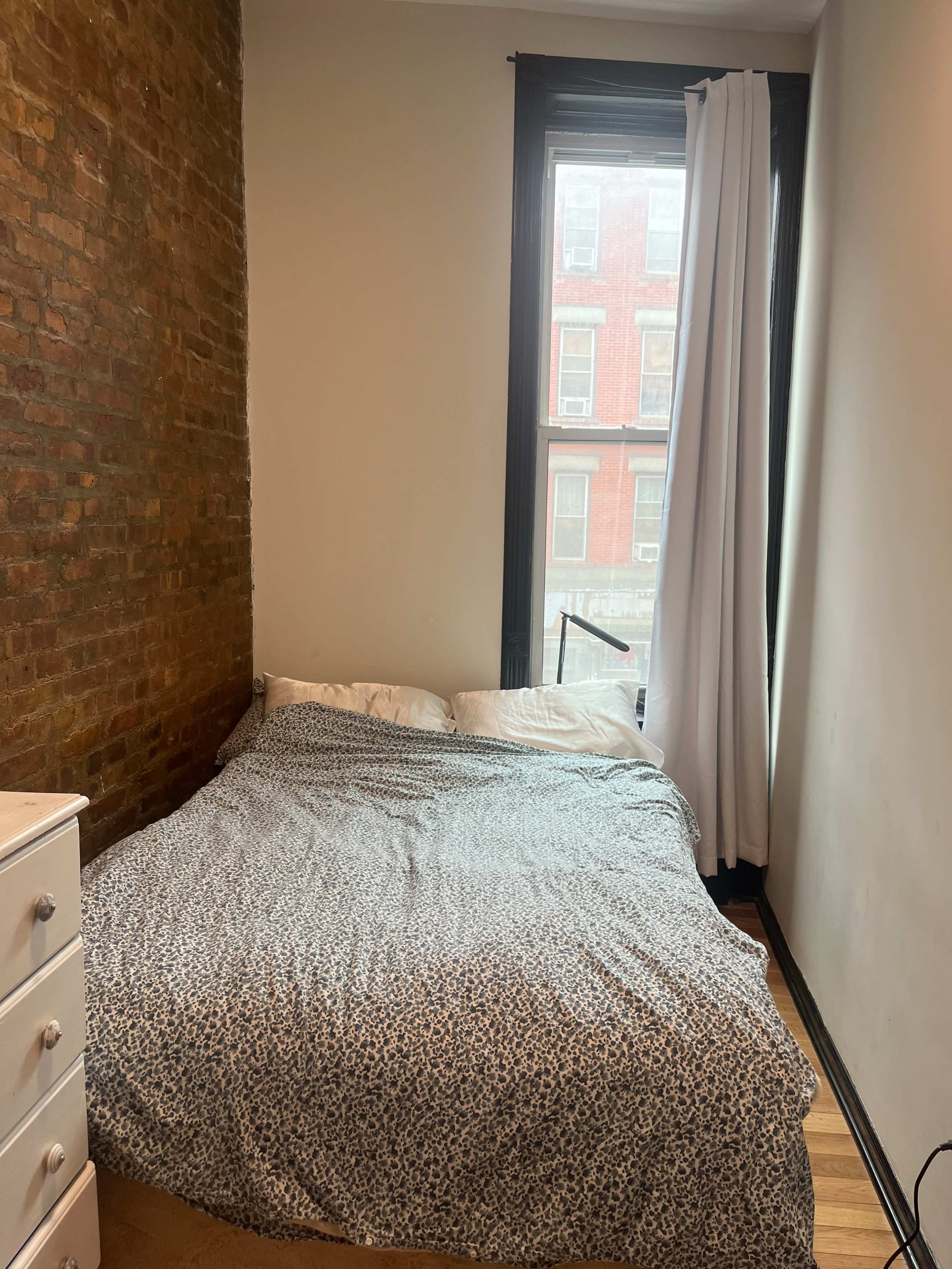 The image shows a simple bedroom with a bed covered in a patterned blanket, a white dresser, and a window with sheer curtains revealing a brick wall and an exterior view.