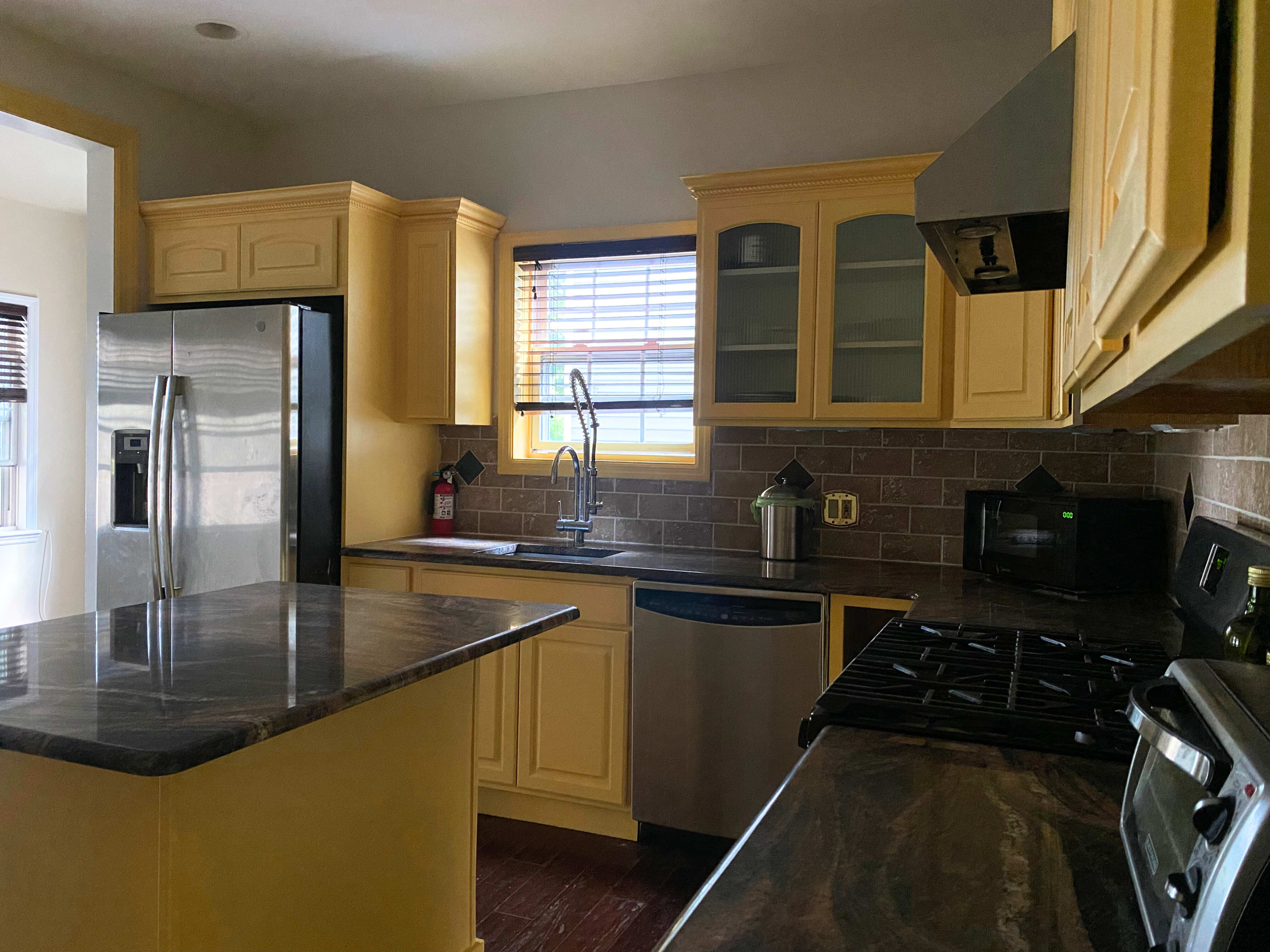 A kitchen with yellow cabinets, a stainless steel refrigerator, a dark countertop, and a window with blinds.