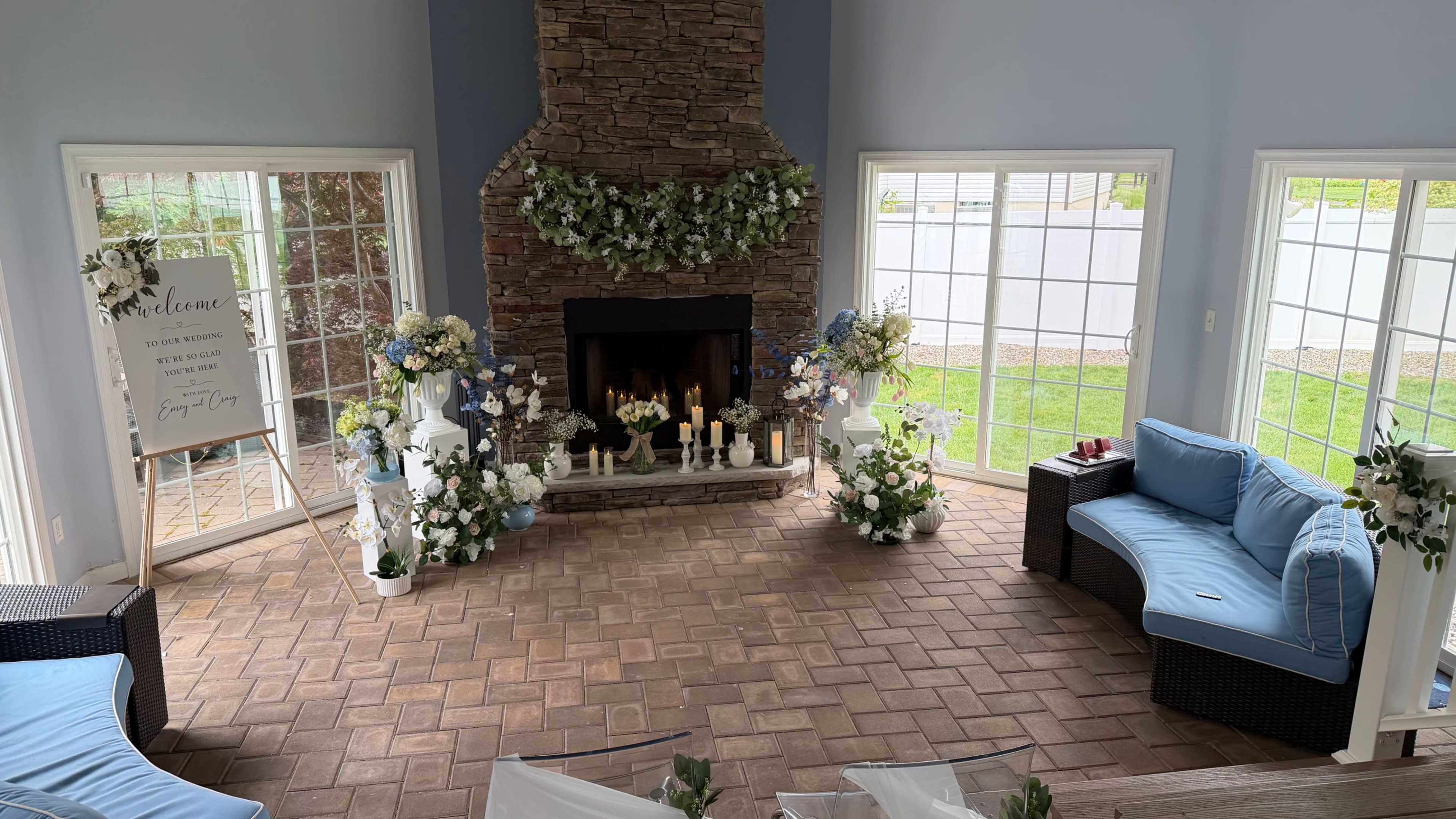 The image shows a spacious room with a stone fireplace, surrounded by flowers and decorative plants, and two blue sofas arranged on either side.