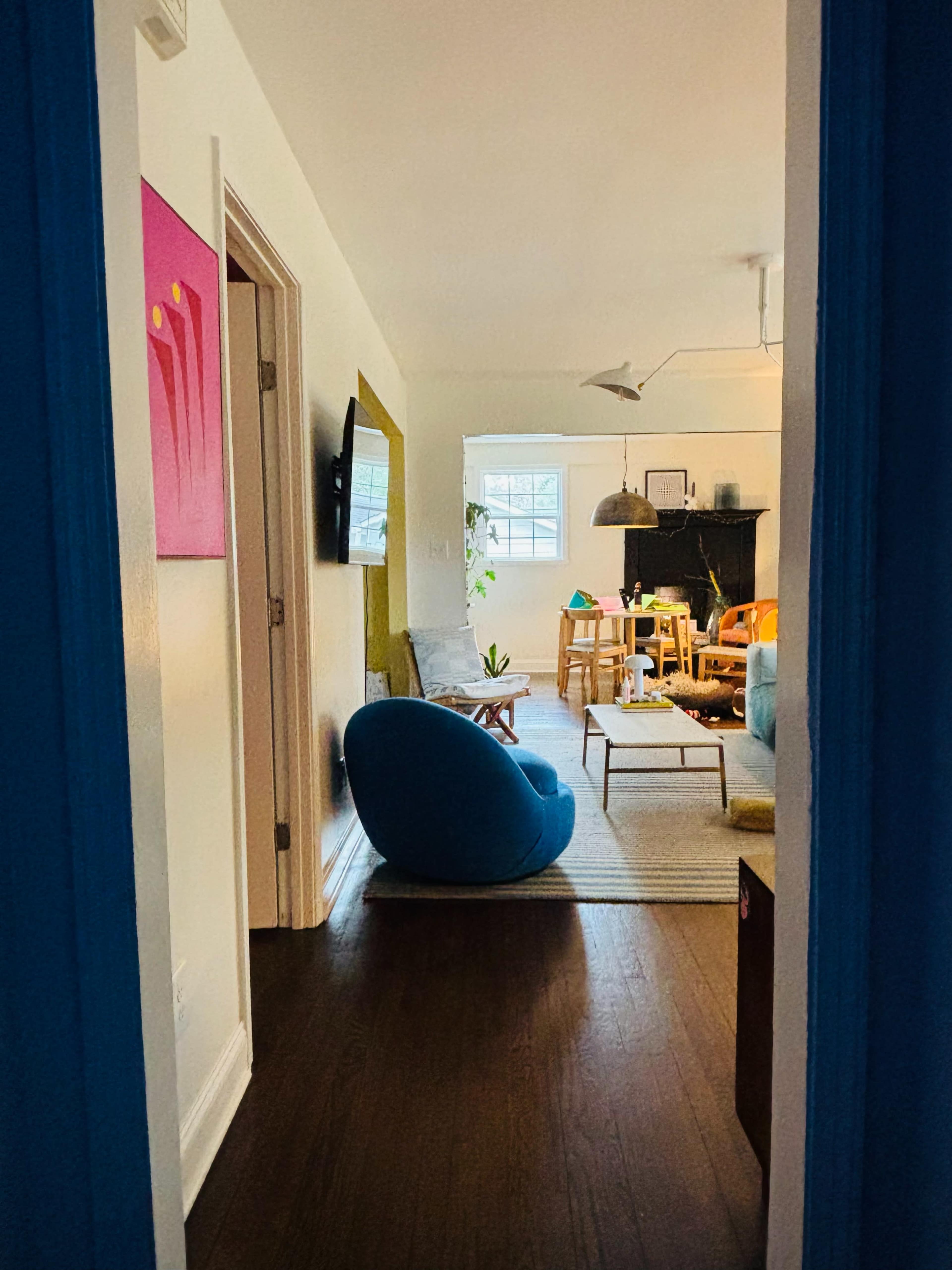 The image shows a view from a doorway into a bright living space featuring a blue bean bag chair, wooden furniture, and a mix of plants and decor.