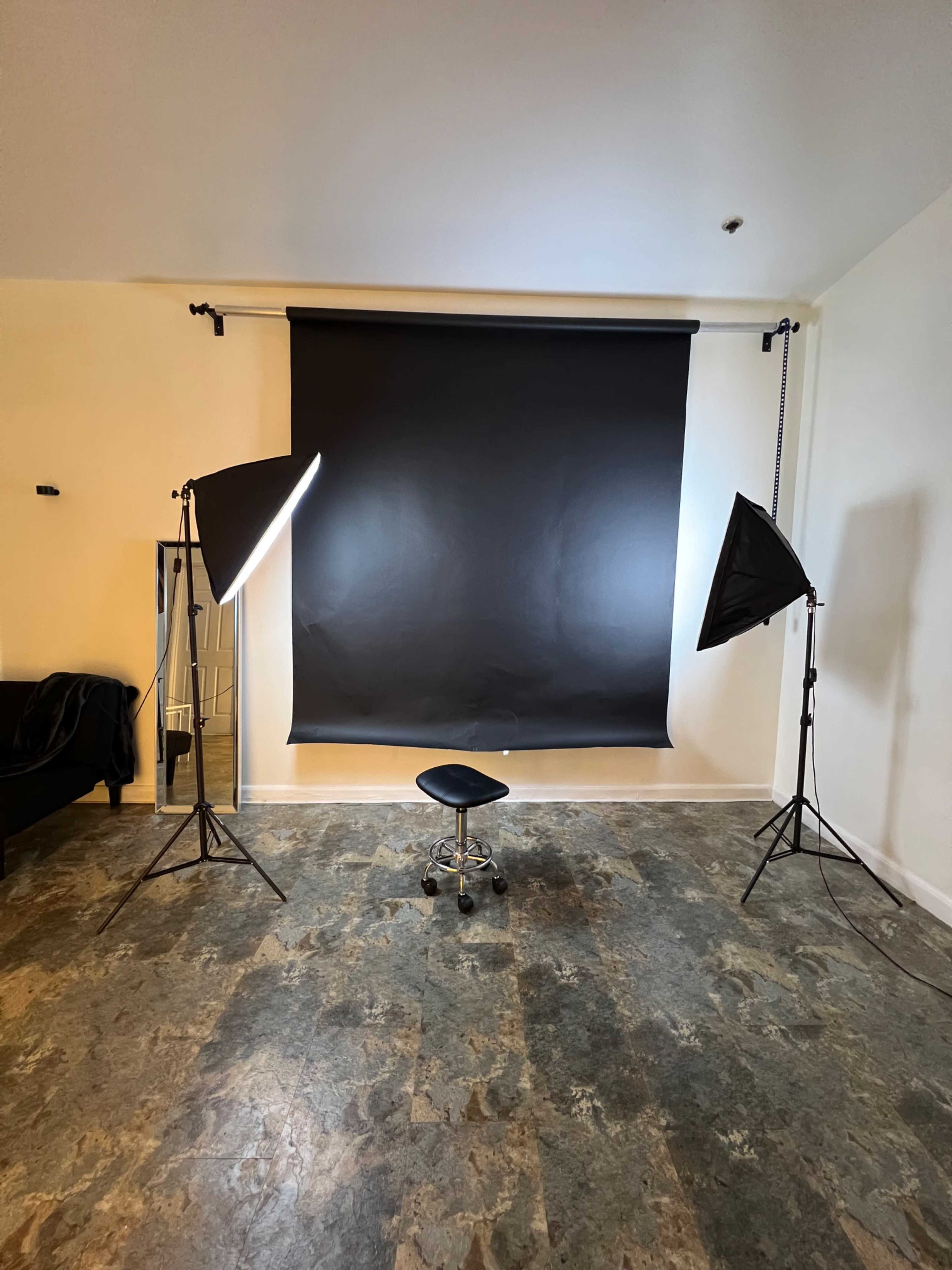 A photography studio setup with two umbrella lights, a black backdrop, and a black stool in the center.