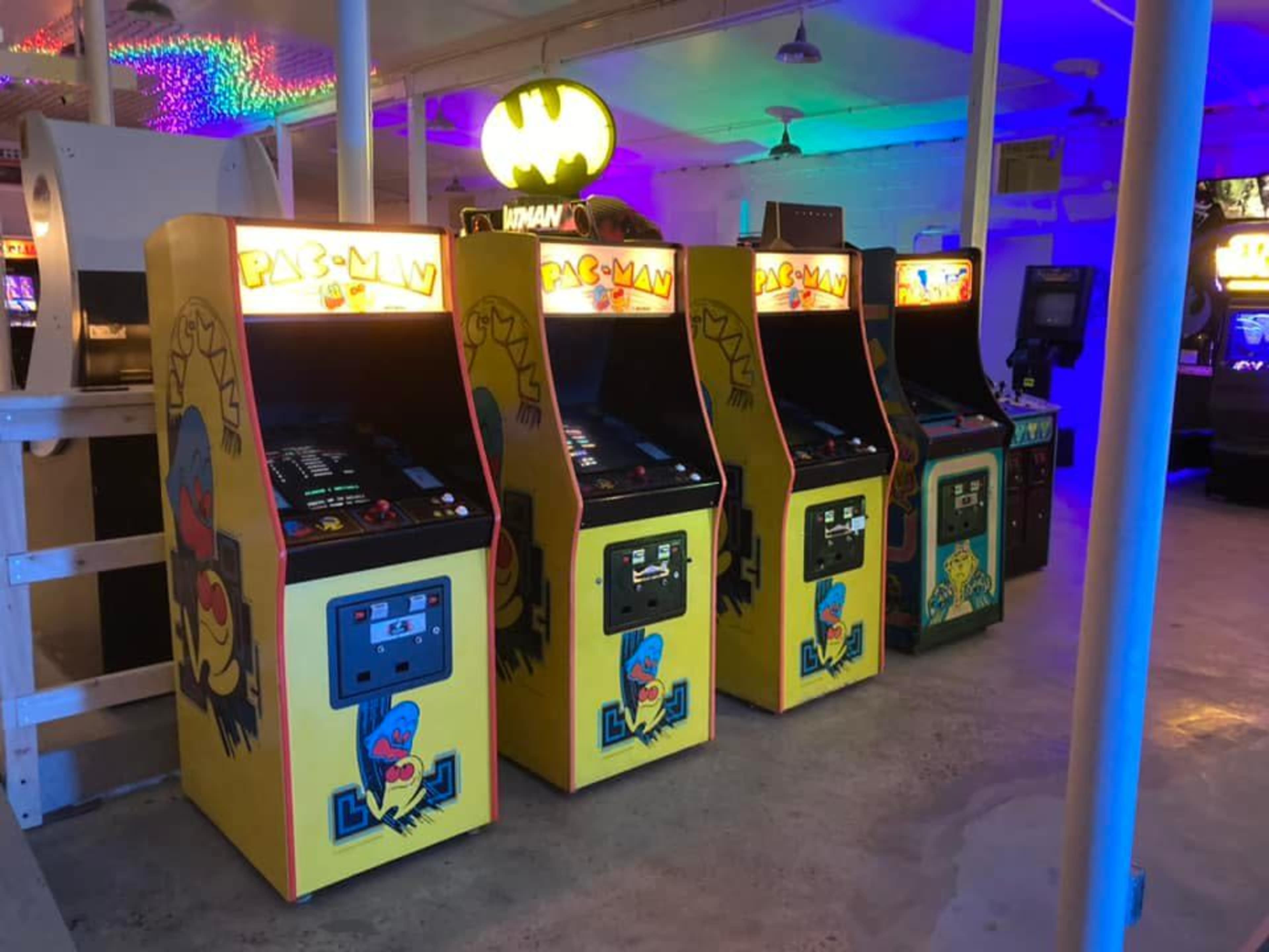 A row of vintage arcade game machines, including Pac-Man, set in a brightly lit arcade with colorful ambient lighting.