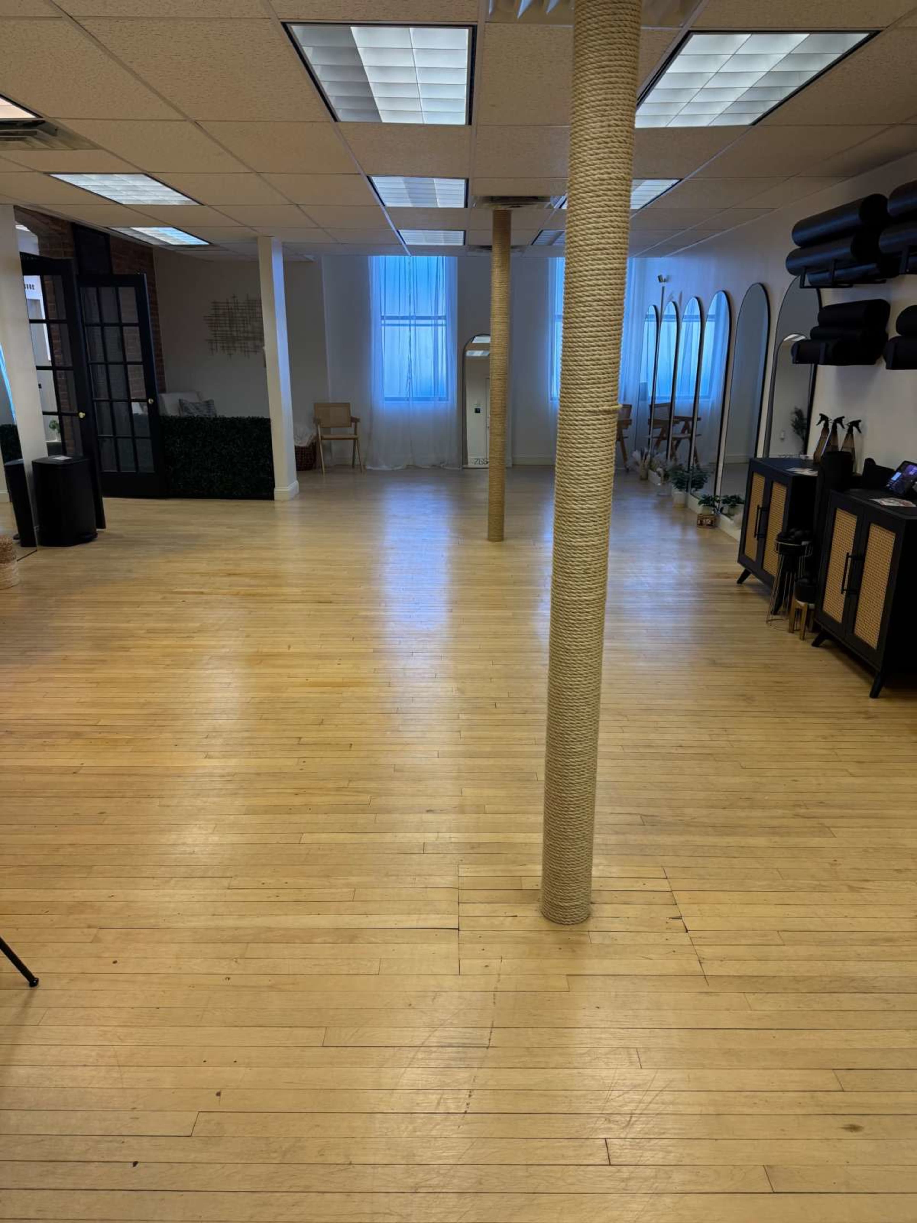 The image shows a spacious room with wooden floors, featuring several pillars, large mirrors along one wall, and a few chairs in the background.