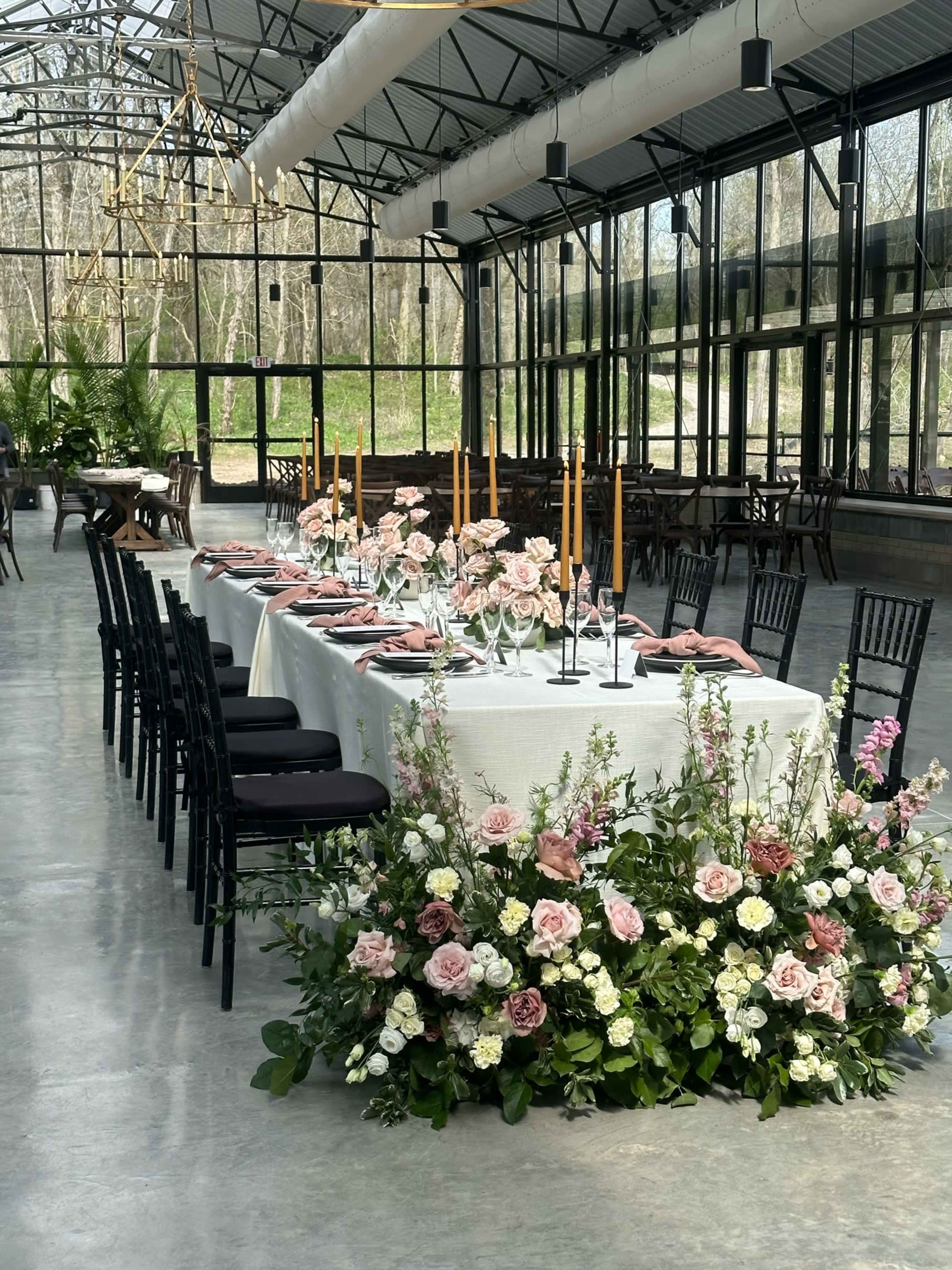 The image features a long dining table set with pink napkins, gold candles, and floral arrangements, surrounded by black chairs in a spacious greenhouse.