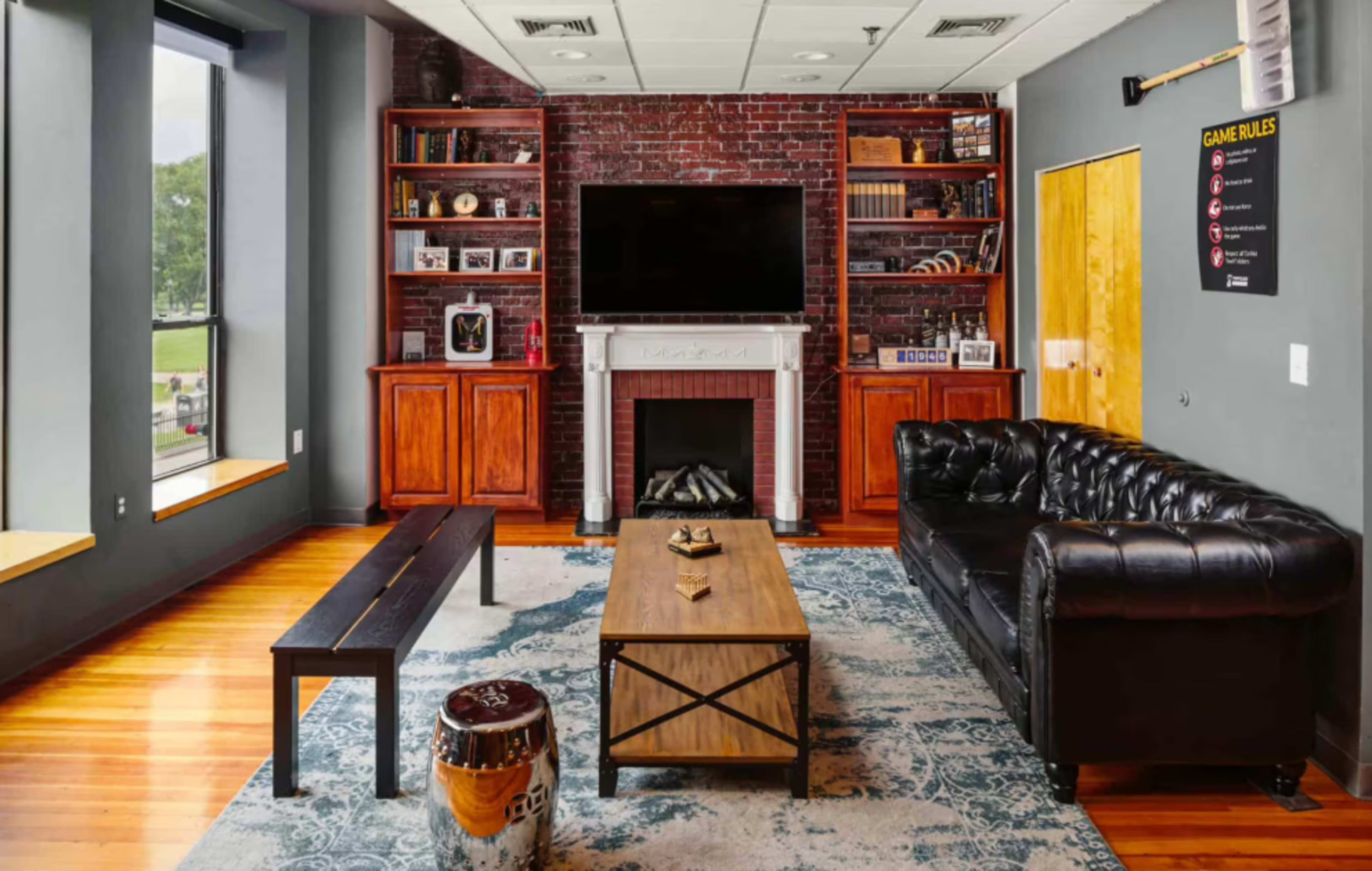 The image depicts a modern lounge area featuring a black leather sofa, a wooden coffee table, a brick accent wall with a television, and shelves with books and decorative items.