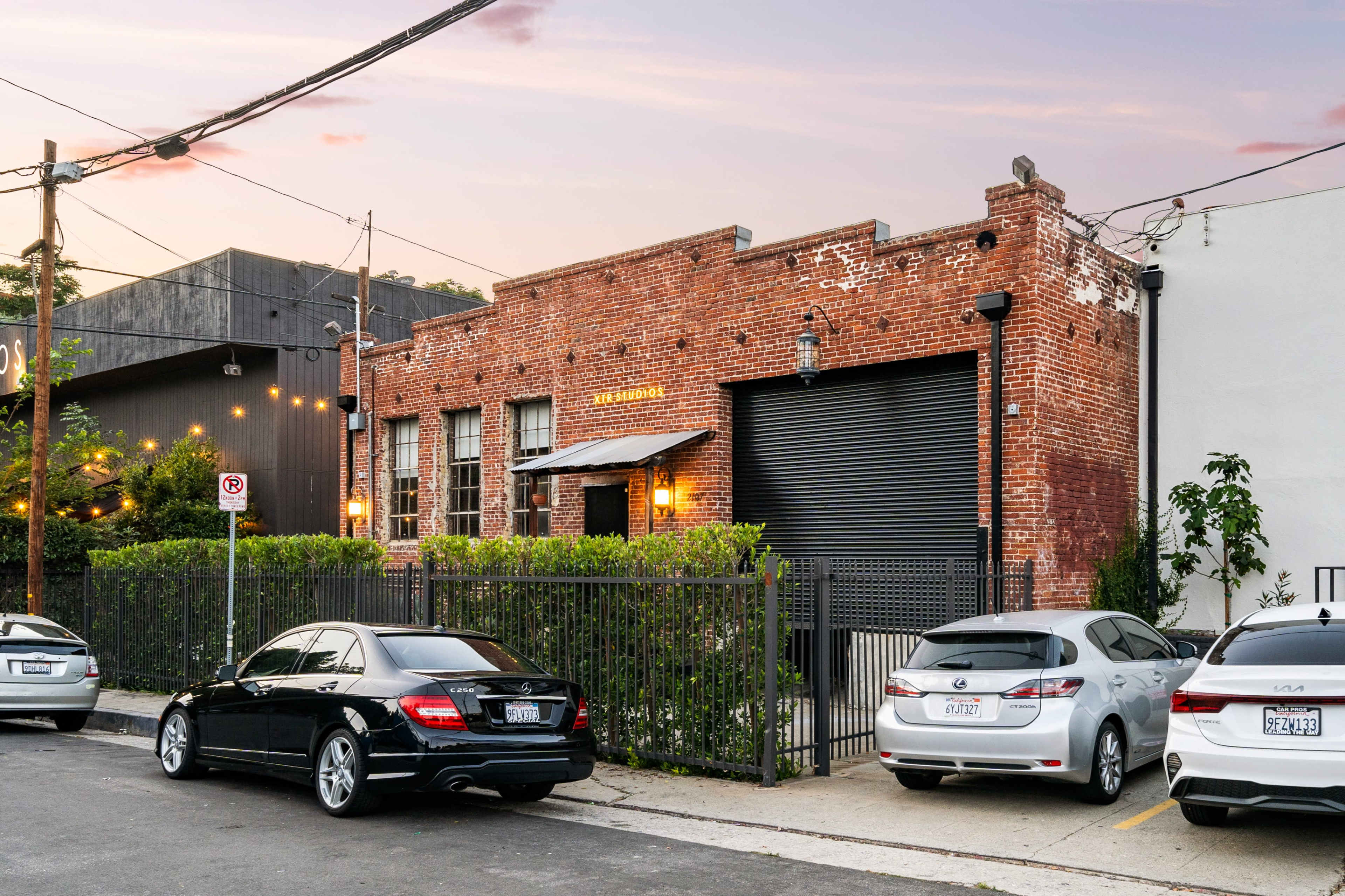 A brick building with a rolled-up black garage door, surrounded by parked cars along the street.