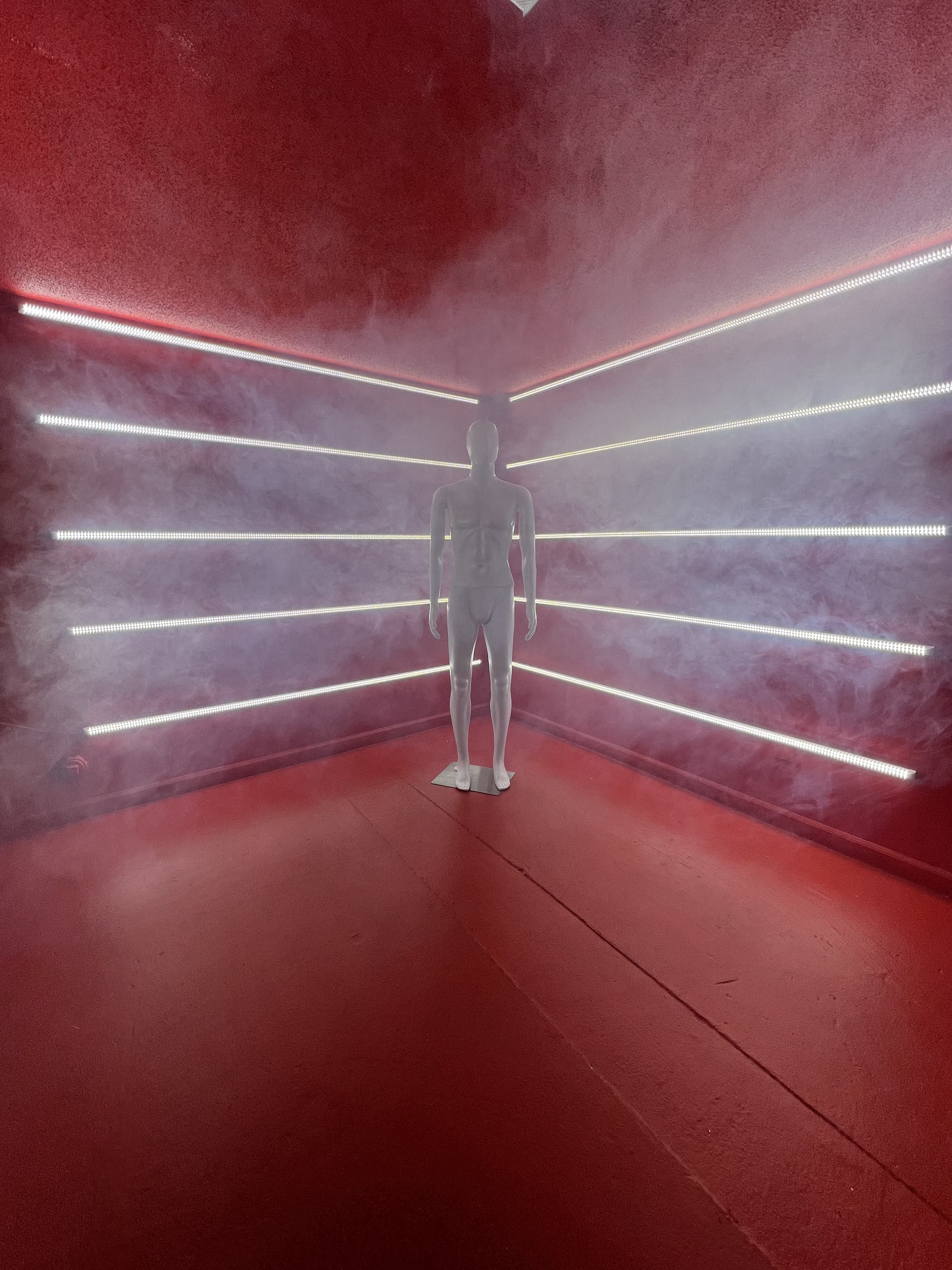 A white mannequin stands in a fog-filled room illuminated by colored LED strips along the walls.