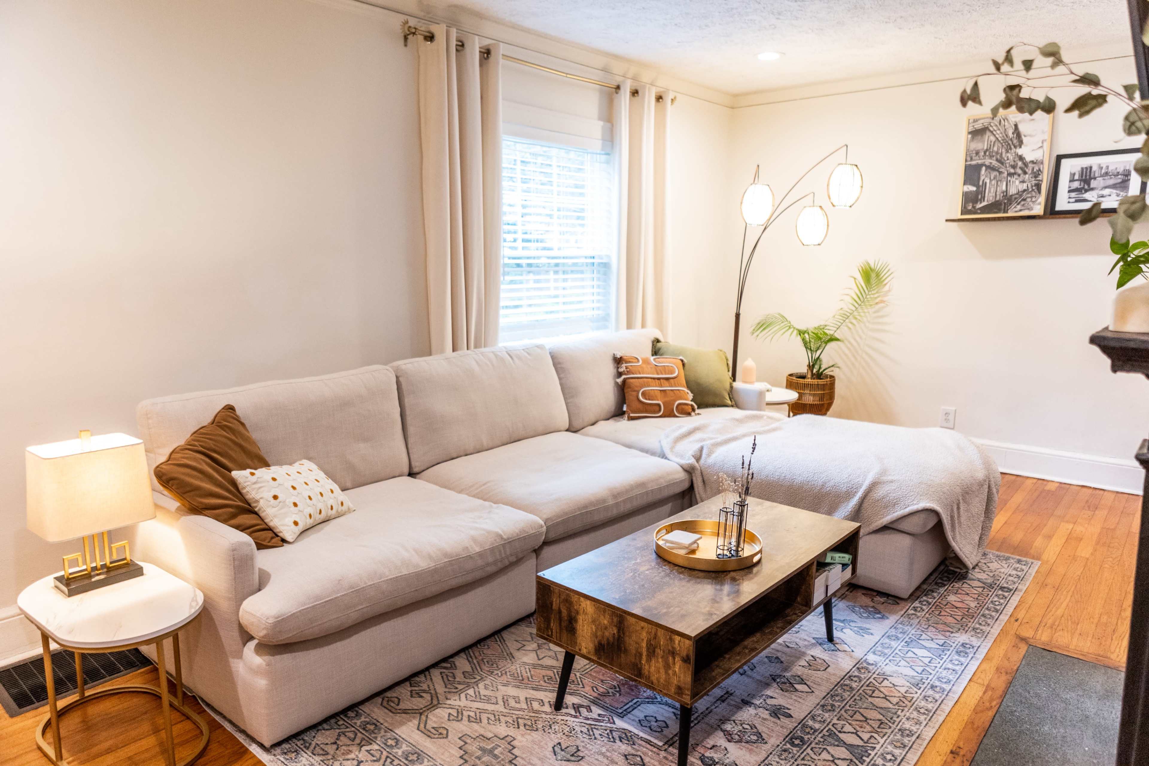 The living room features a beige sectional sofa, a wooden coffee table, a floor lamp with three curved arms, and a small plant beside a window.