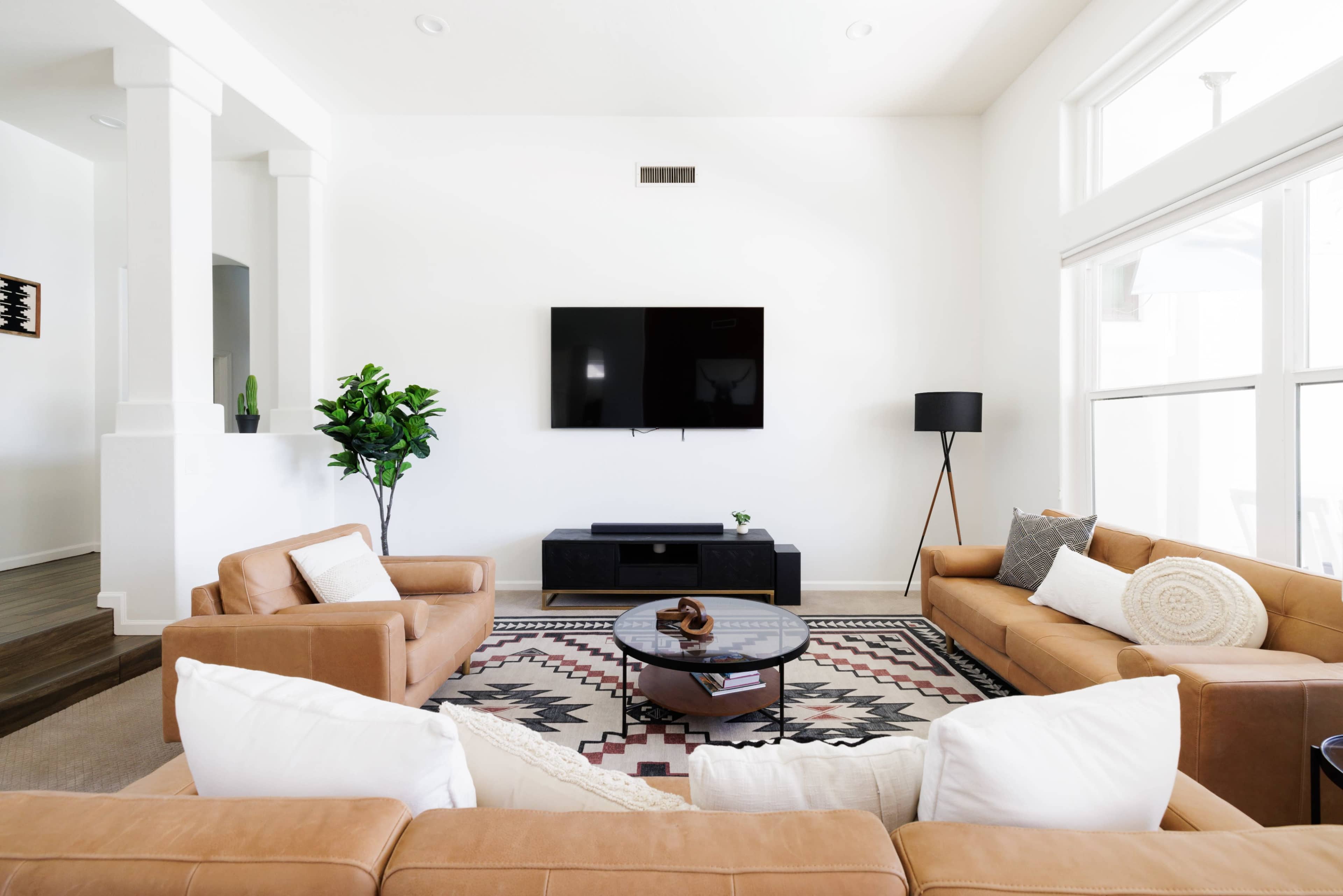 A modern living room features two tan leather sofas, a circular coffee table, and a flat-screen TV mounted on a white wall, with large windows allowing natural light.