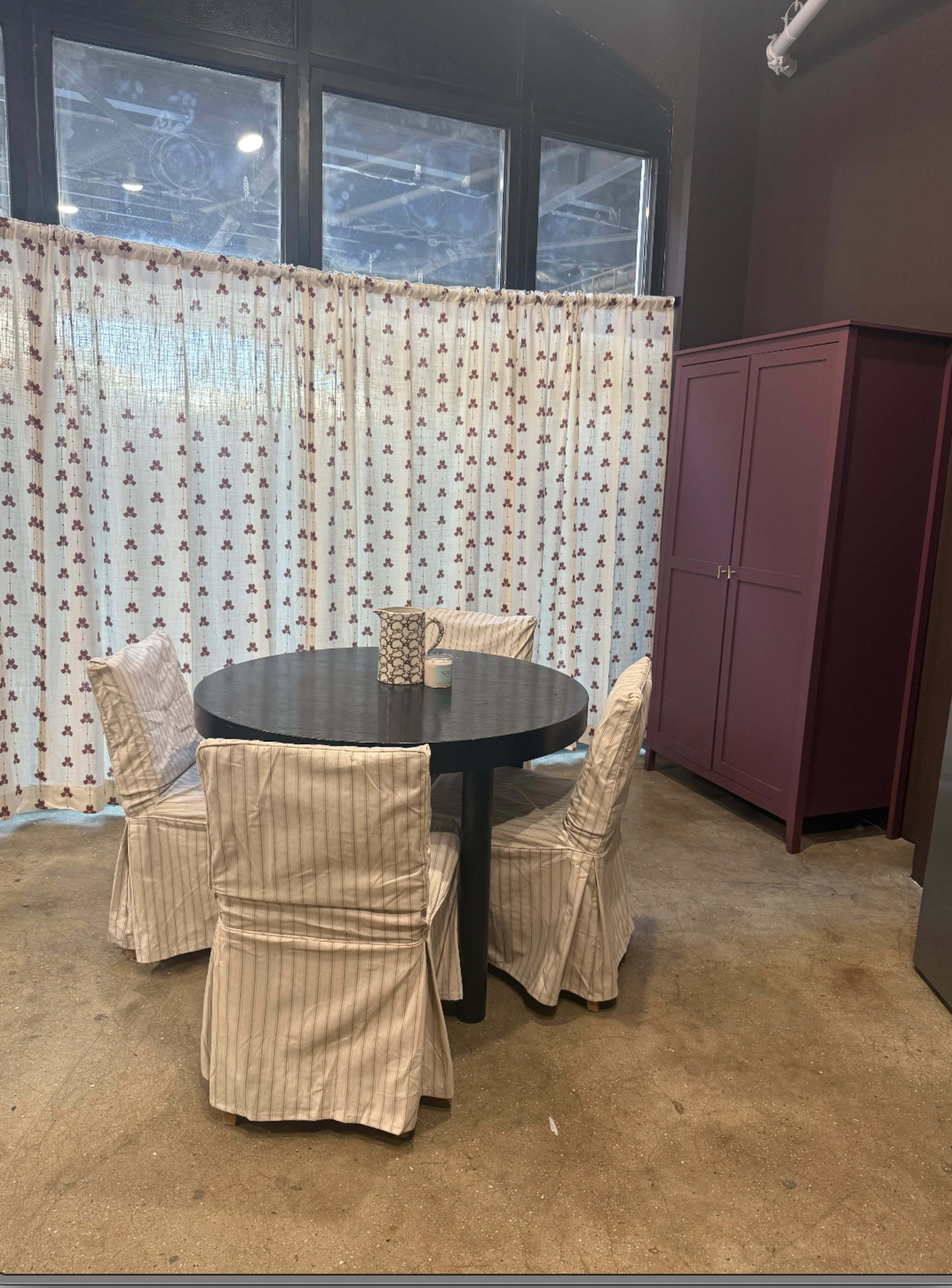 The image shows a round black dining table surrounded by four patterned chairs, with a purple cabinet against a wall and sheer curtains in the background.