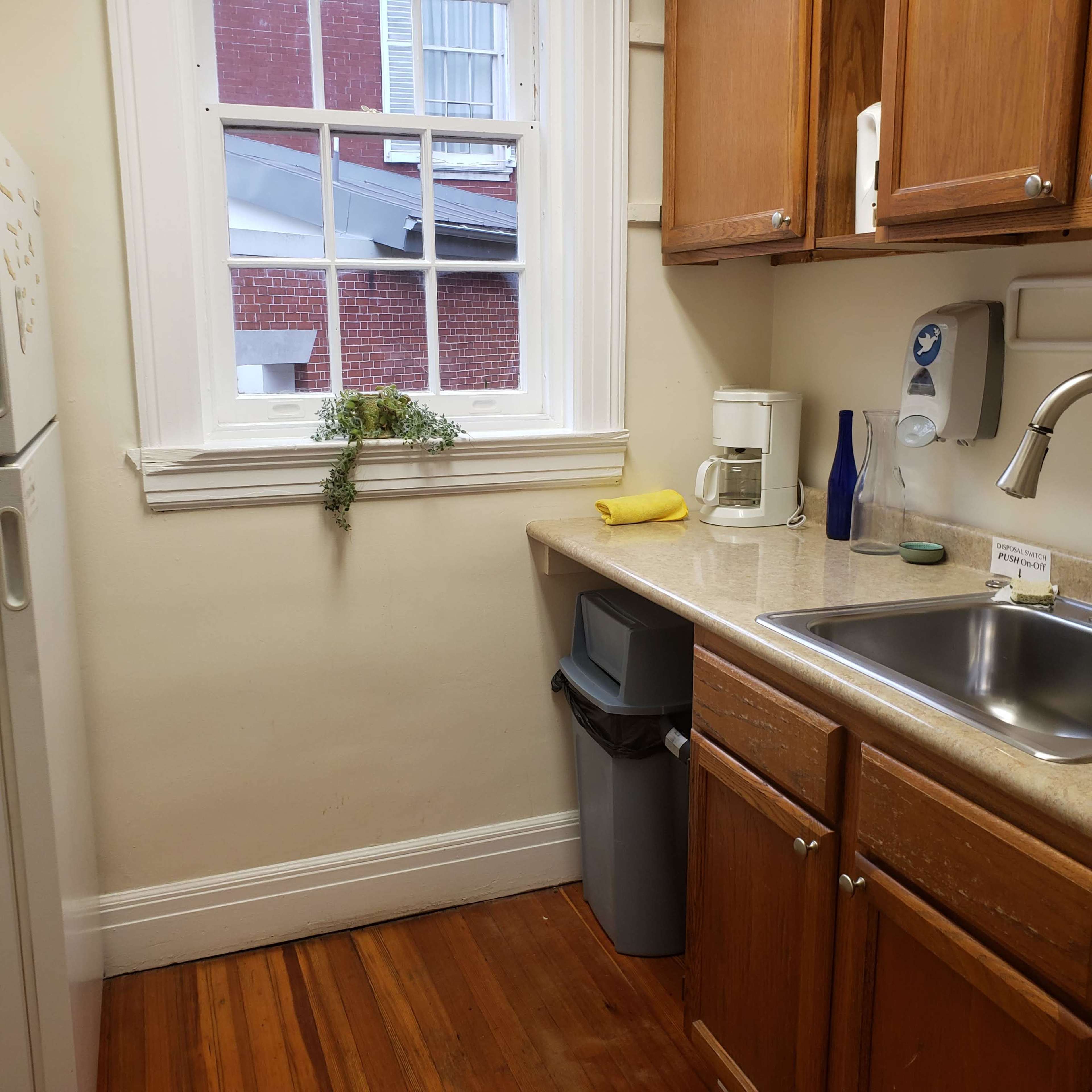 A small kitchen corner features a sink, a coffee maker, a trash bin, and wooden cabinets, with a window letting in natural light.